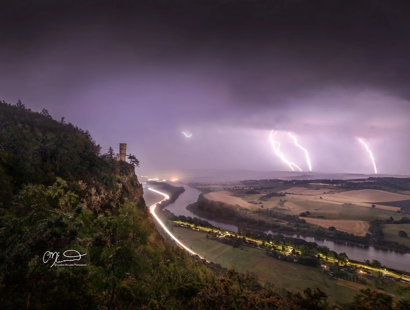 Scotland, Central Highlands, Kinnoull Hill. Nighttime landscape showing a river winding through a valley with light trails from moving vehicles, a hill on the left with a tower or castle ruin, and multiple lightning bolts striking in the cloudy sky.