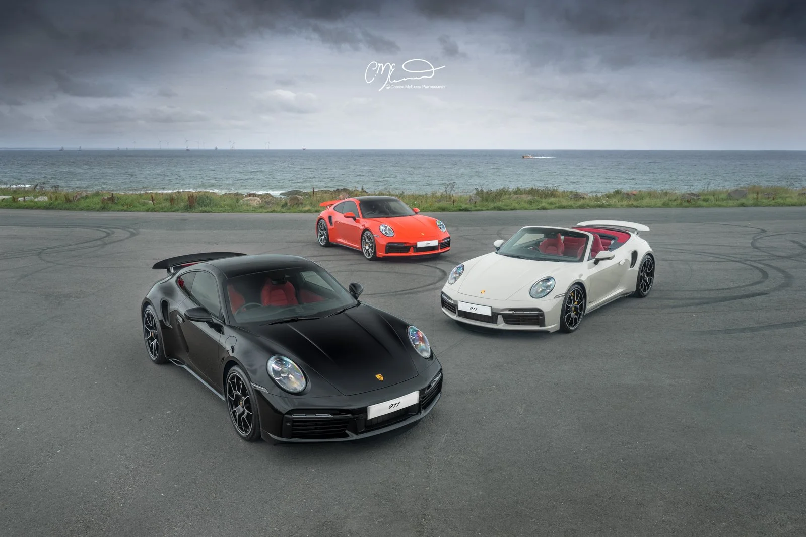 Three Porsche 911 Turbo S, 992 cars parked on an empty asphalt area near the coast during cloudy weather, with the ocean and wind turbines visible in the background.
