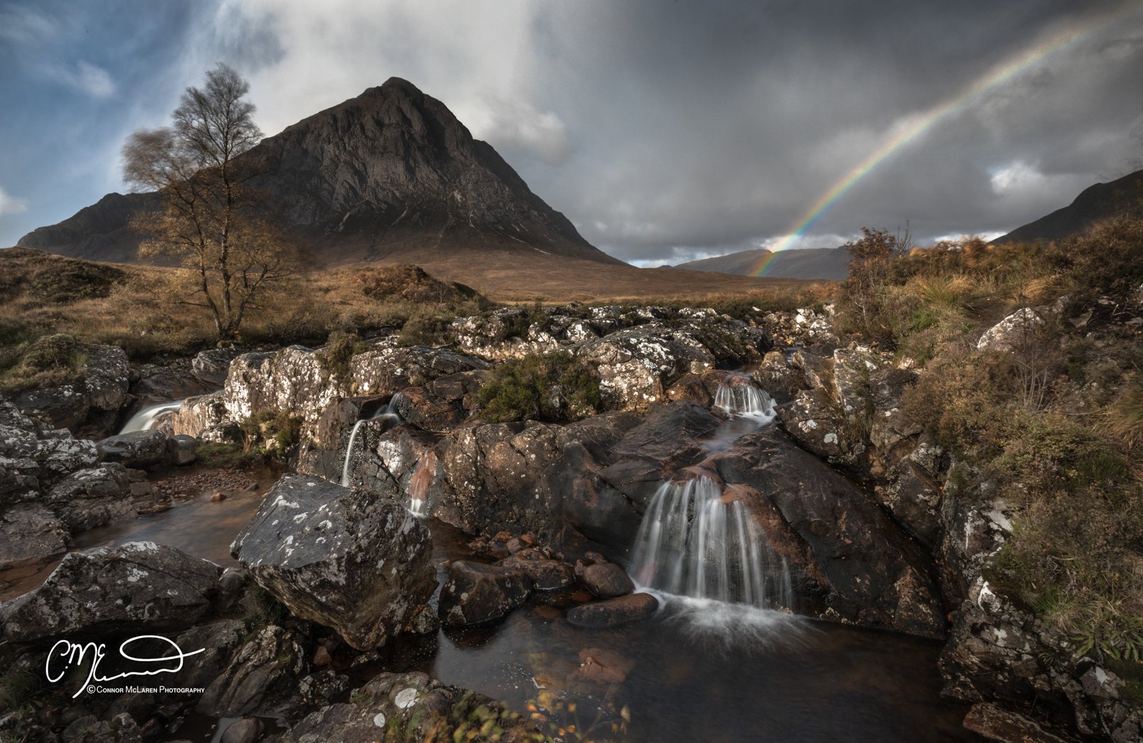 Glen Coe and Glen Etive, Western Highlands, Scotland. Mountain landscape with a river flowing over rocks, a rainbow in the sky, and storm clouds above.