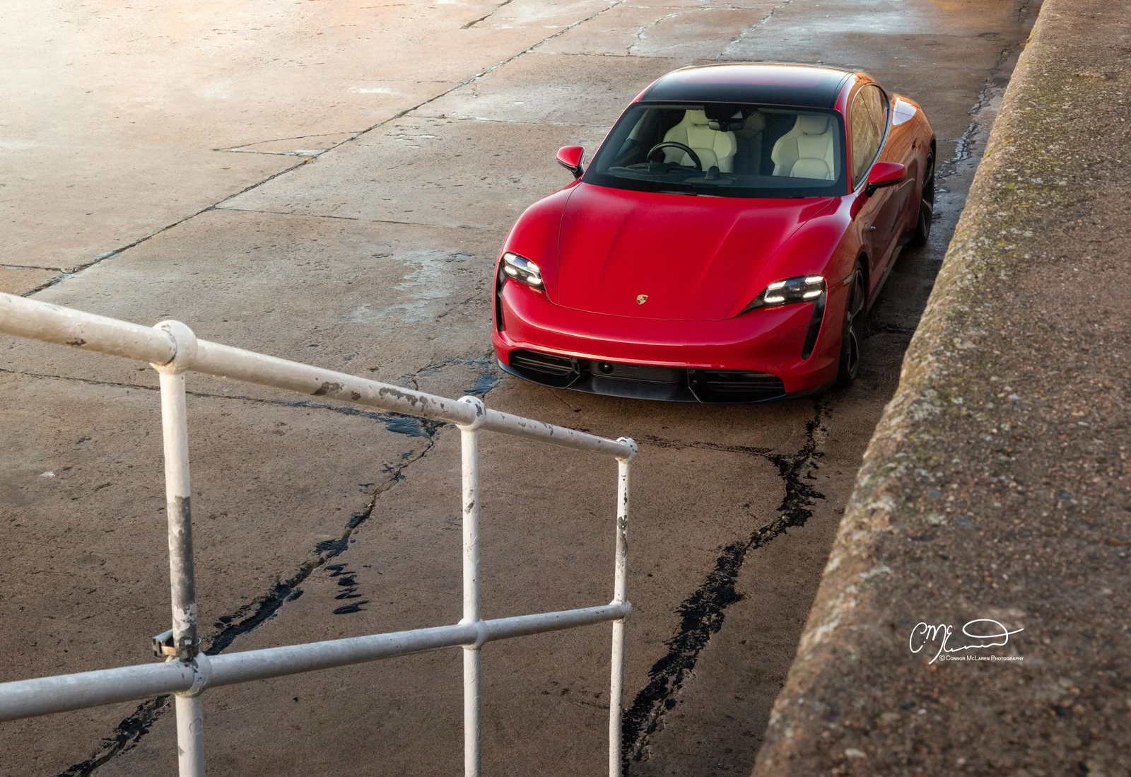 Red Porsche Taycan sports car parked on a cracked concrete surface next to a rusty white metal railing and a weathered stone wall.