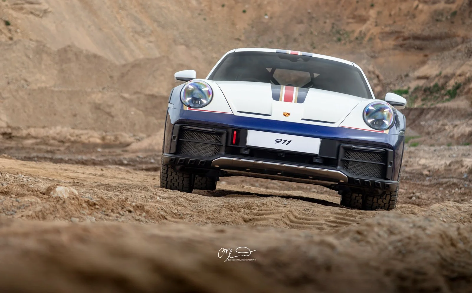 A whitePorsche 911 Dakar Roughroads sports car with racing stripes driving on a dirt road in a desert landscape.