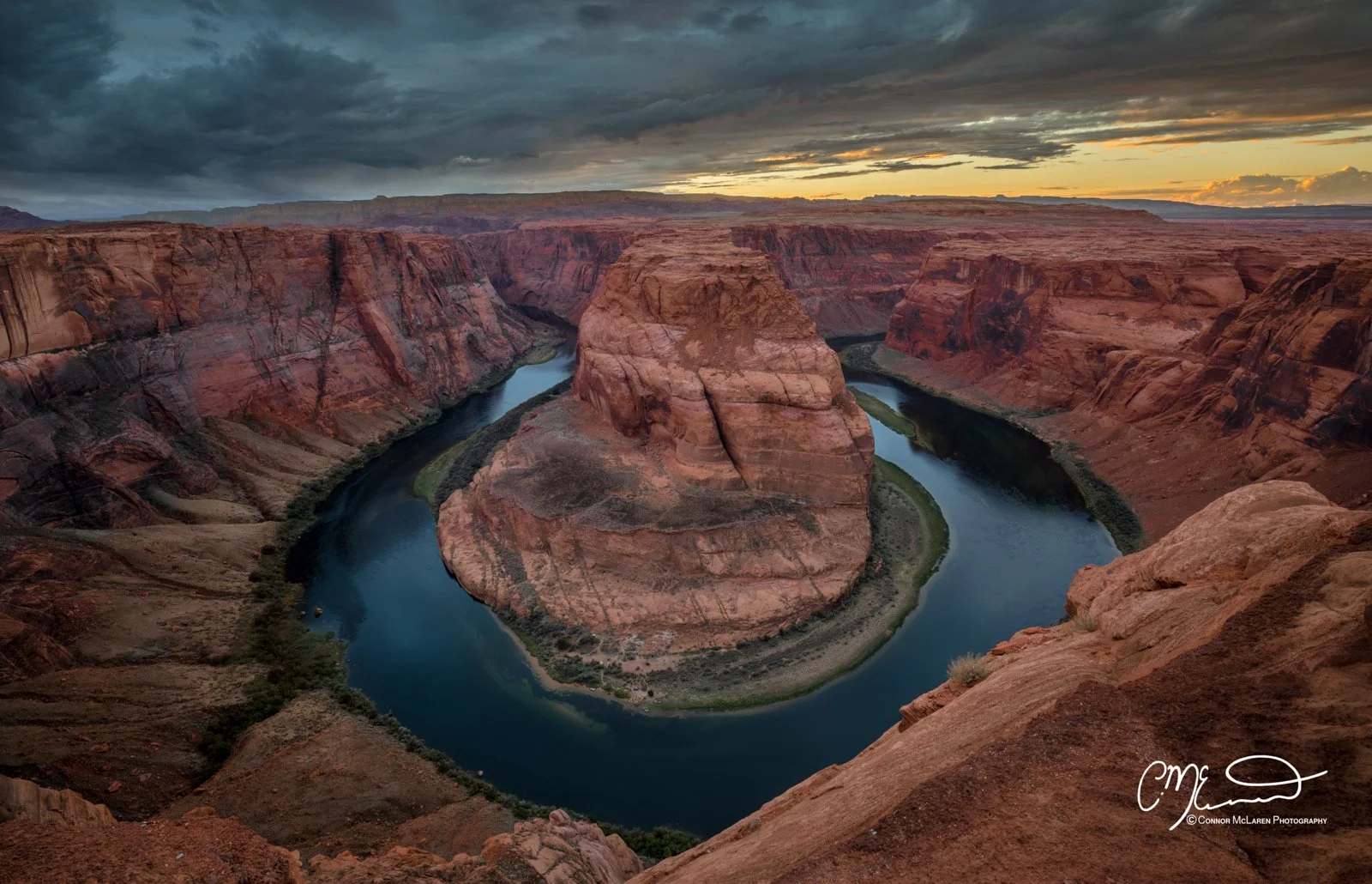 Sunset over the Horseshoe Bend, a 1,500-foot-wide meander of the Colorado River in Arizona, with dramatic red rock formations and cloudy sky.