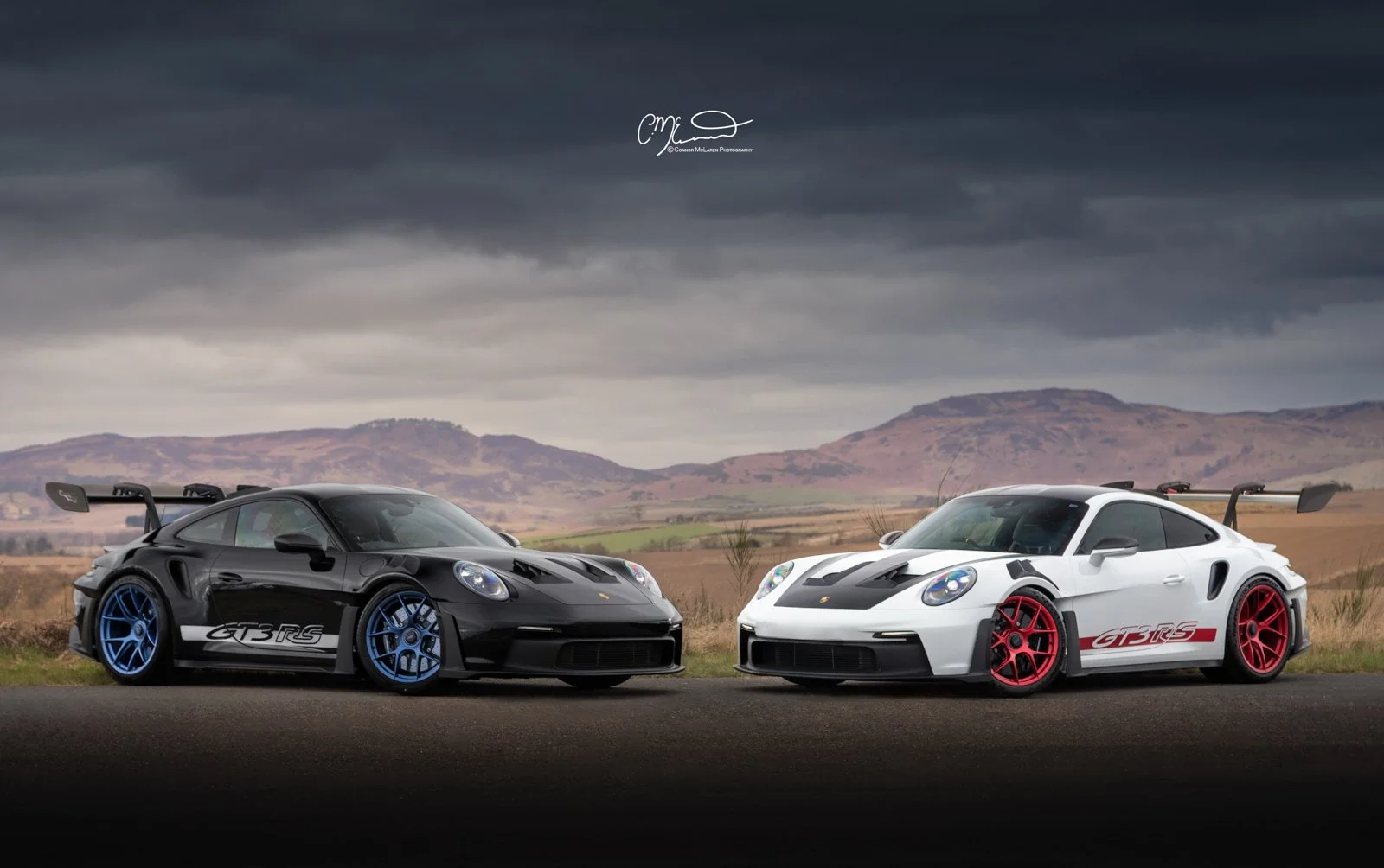 Two Porsche 911 GT3 RS cars, one black with blue accents and one white with red accents, parked on a road with mountainous landscape and dark cloudy sky in the background.