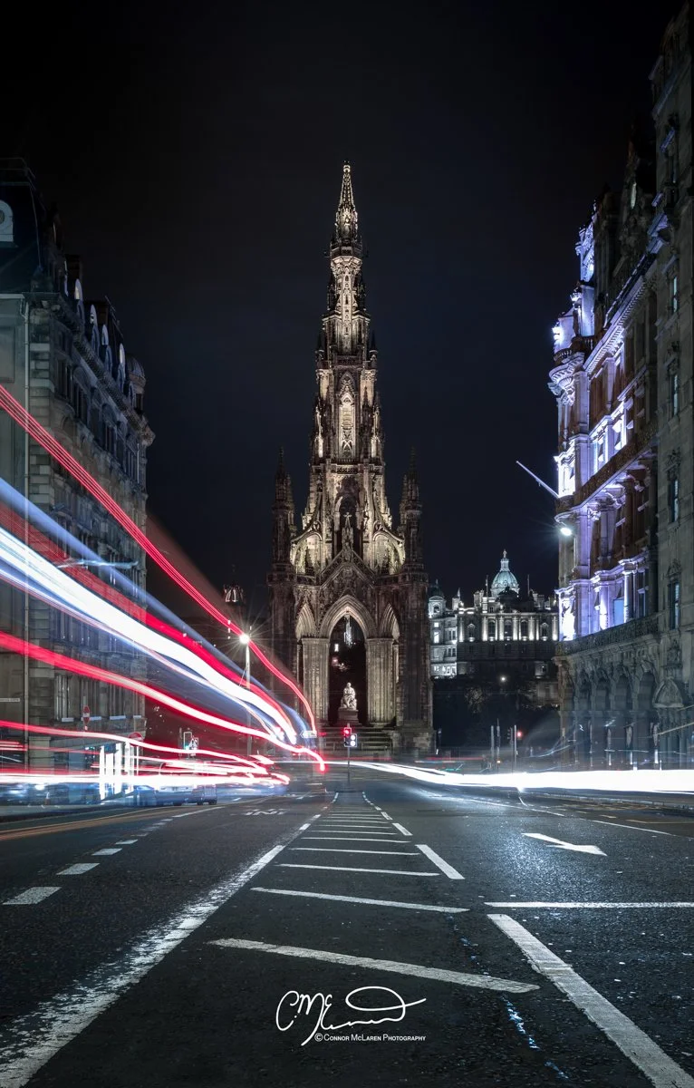 Scottish Lowlands, Scotland,  Edinburgh. Nighttime city street with historic cathedral in the background, light trails from moving vehicles, and illuminated buildings on both sides of the street.