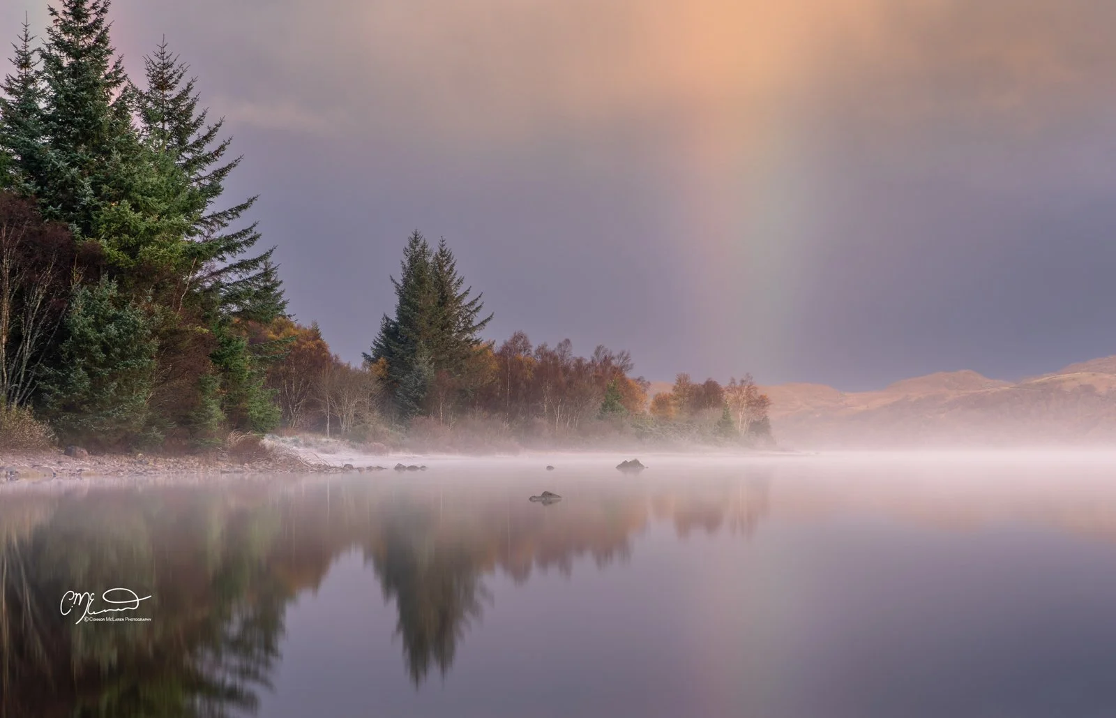 Scotland, north coast 500, NC500, Loch Maree. Calm river scene with mist and colorful trees, hills in the background, and a faint rainbow in the sky at dawn or dusk.