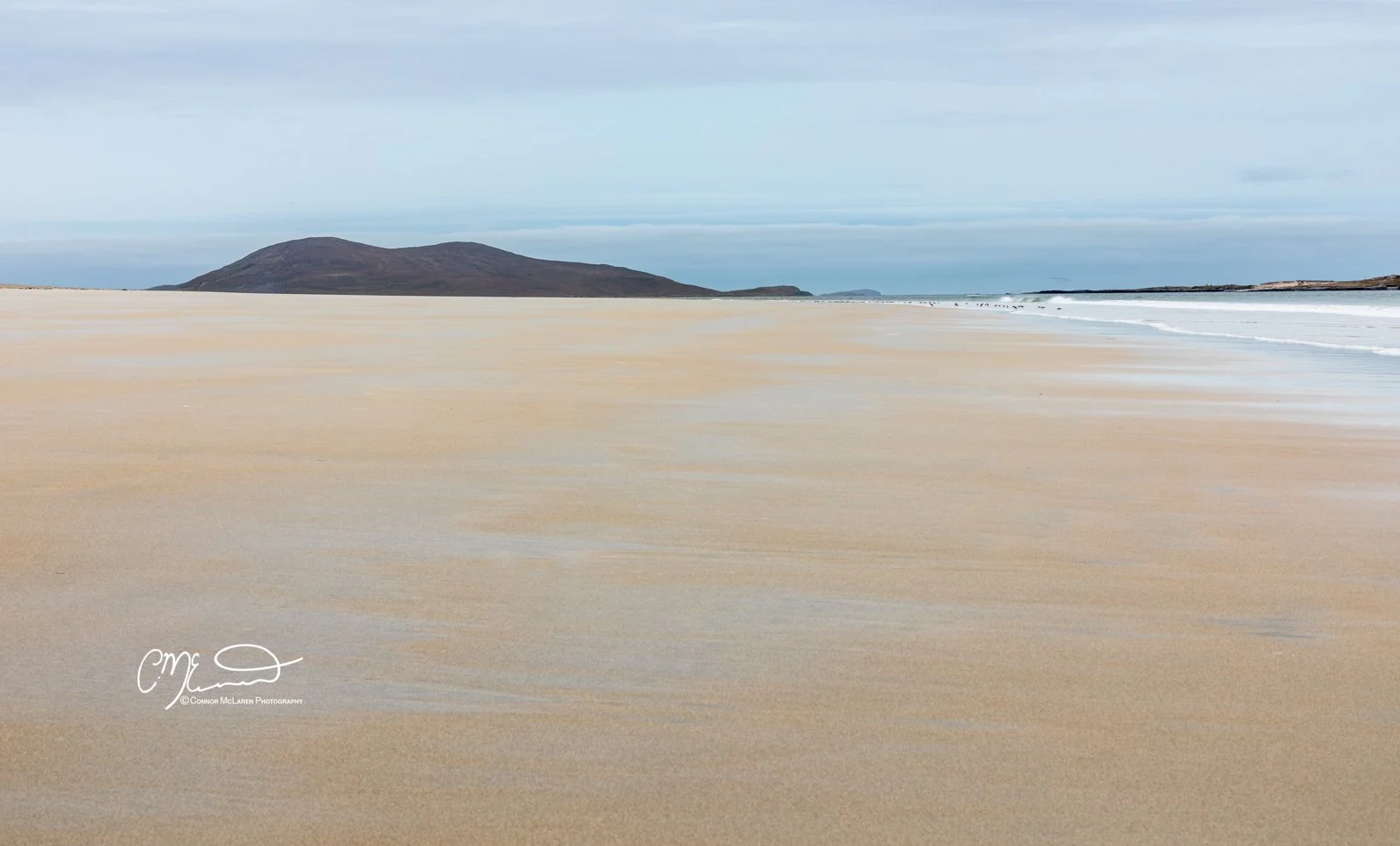 Scotland, Lewis Harris & Outer Hebrides, Luskentyre Beach. Wide beach with golden sand, calm ocean waves, and a distant dark mountain under a cloudy sky