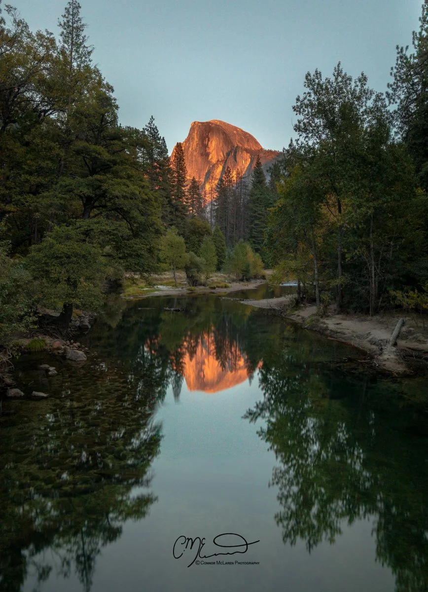 Bucket list locations, USA Yosemite National Park. Sunset lighting up a mountain peak over a forest reflected in a calm river surrounded by trees.