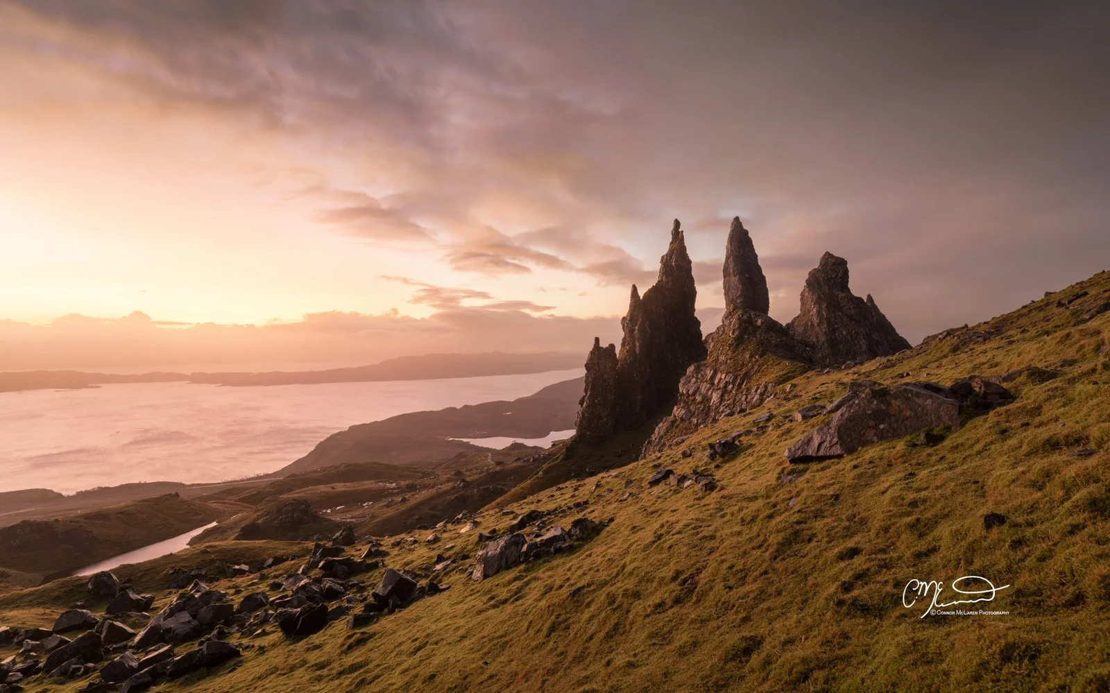 Bucket list locations, Scotland, Isle of Skye, old man of storr. Sunset over a landscape with rugged green hills and three prominent tall rock formations, with a body of water and distant mountains in the background.