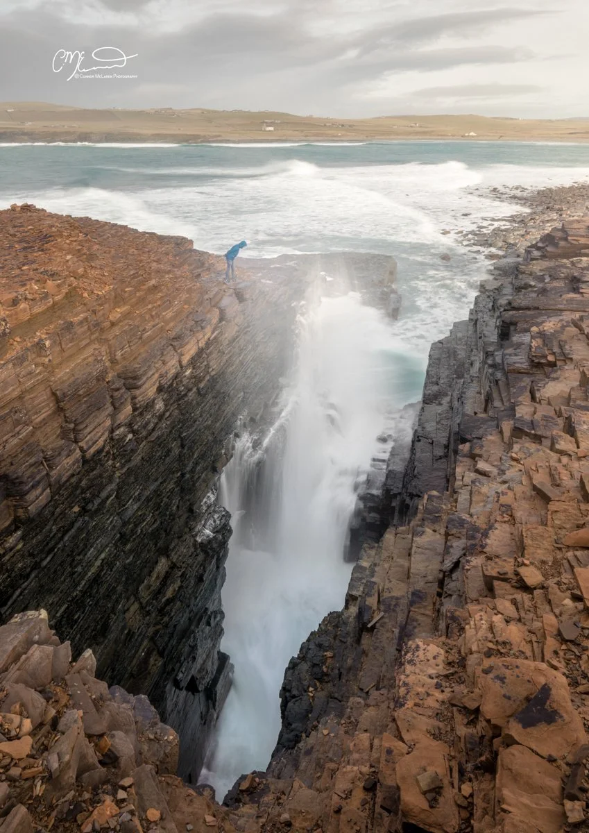 Orkney, Scotland. Person standing on rocky cliff overlooking a waterfall flowing into the ocean, with hills and cloudy sky in the background.