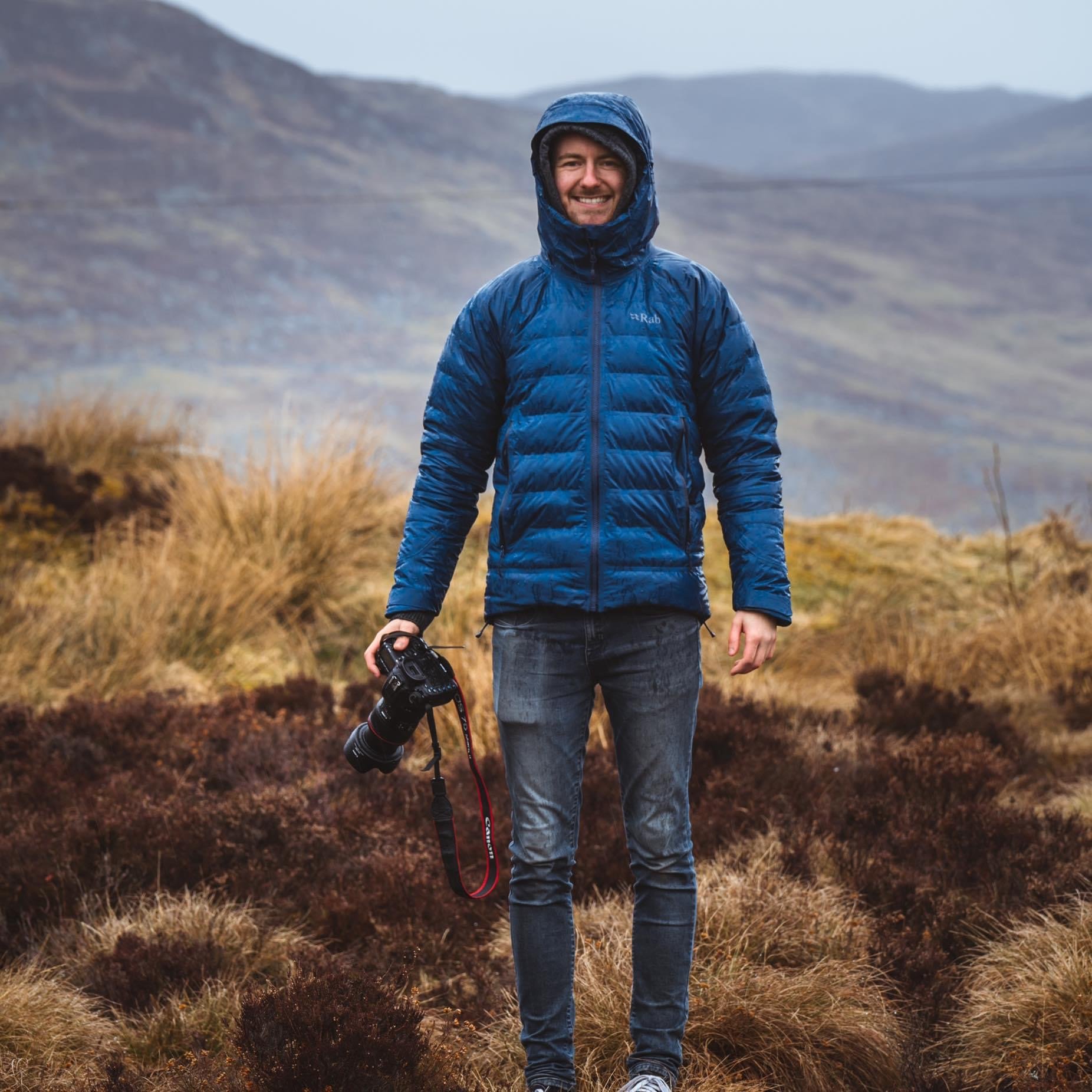 Connor McLaren Photography, Photographer. Connor McLaren in a mountainous area with brownish grass, wearing a blue insulated jacket with the hood up, holding a camera in his right hand, smiling at the camera.