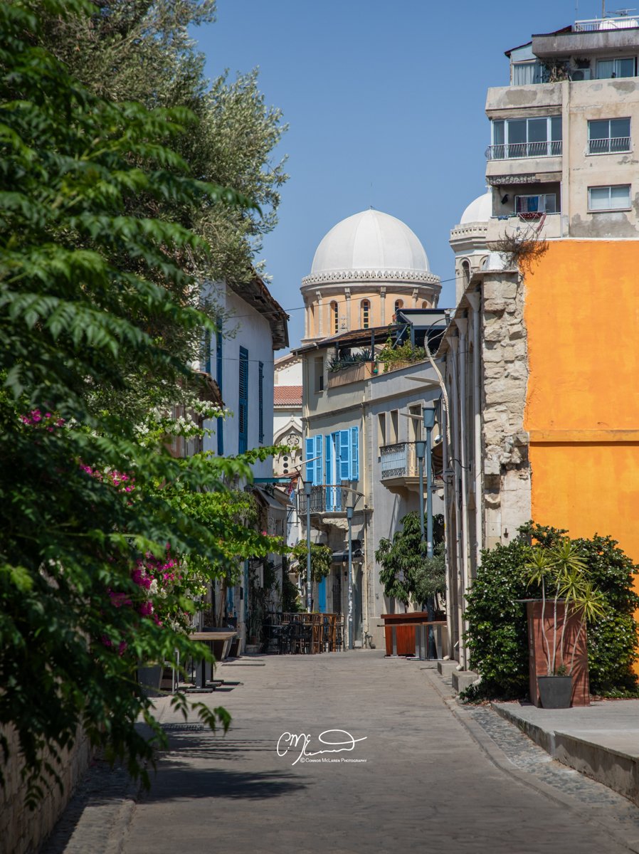 Limassol, Cyprus. A narrow street in a Mediterranean city with vibrant buildings, greenery, and a large domed structure in the background.
