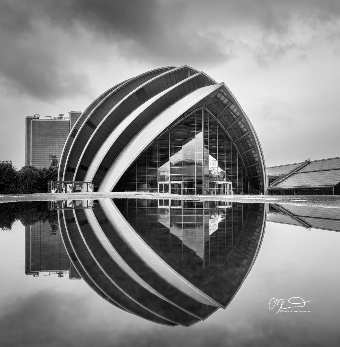 Scotland, Glasgow SECC Armadillo. Modern building with curved roof and glass facade, reflected in a pool of water, under cloudy sky.