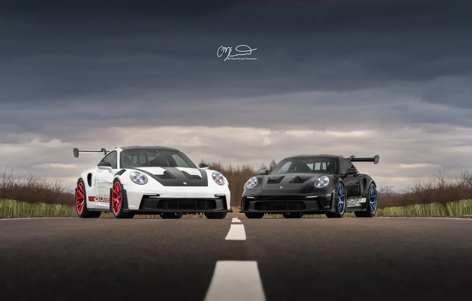 Two Porsche 911 GT3 RS sports cars parked facing each other on an empty road under a cloudy sky, one in white with red wheels and the other in black with blue wheels.