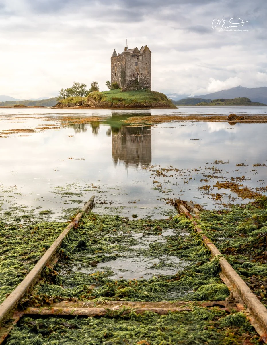 Castle Stalker, Western Highlands, Scotland. An old, rusty railroad track covered in green algae leading to a small island with a medieval castle, reflected in calm water under a cloudy sky.