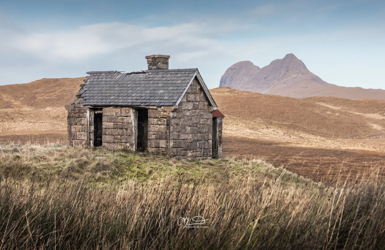 Scotland, north coast 500, NC500, Assynt. Suilven. An old stone house in a vast, barren landscape with brown grassy hills and two mountains in the distance under a partly cloudy sky.