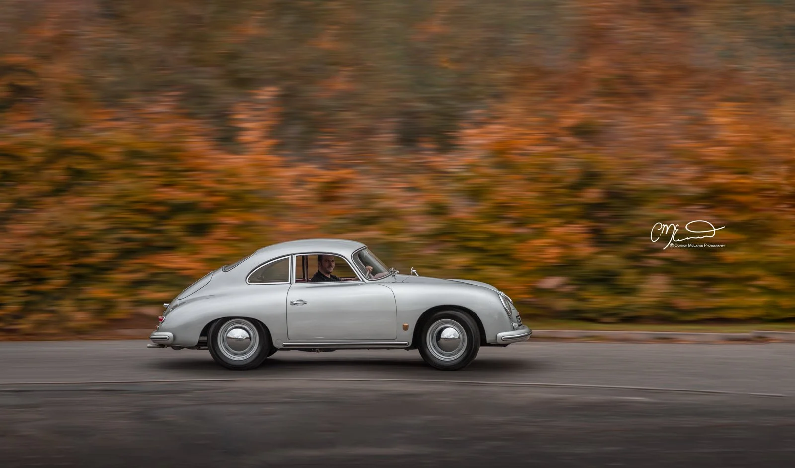 Porsche 356. A vintage silver car driving on a road with a blurred autumn-colored wooded background.