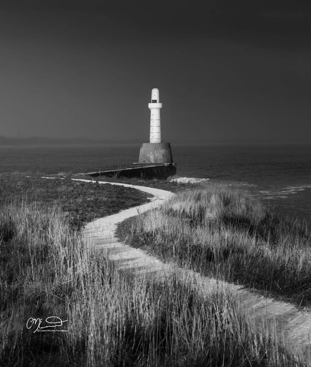 Scotland, Aberdeen Lighthouse. A black and white photo of a lighthouse on a rocky shoreline, with a winding path leading to it through tall grass.