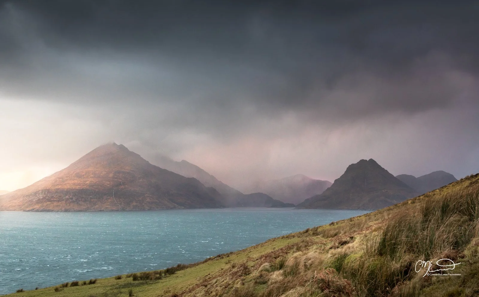 Scotland, Isle of Skye, Elgol. A scenic landscape featuring a large body of water with mountains in the background under a dark, stormy sky and grassy hills in the foreground.