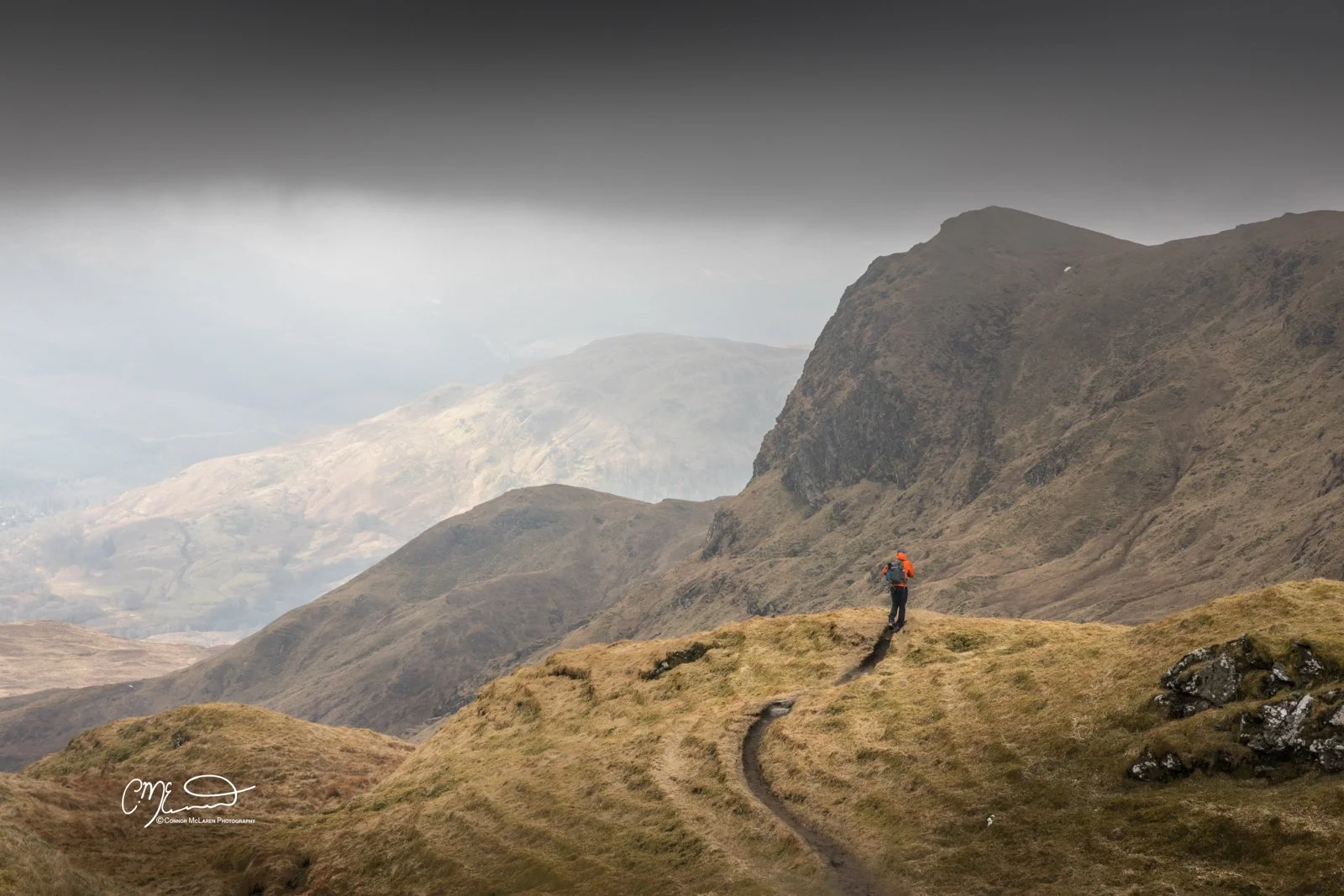 Scottish Central Highlands, Scotland, Perthshire munro. A lone hiker in an orange jacket walking on a grassy mountain trail during daytime with misty mountains in the background.