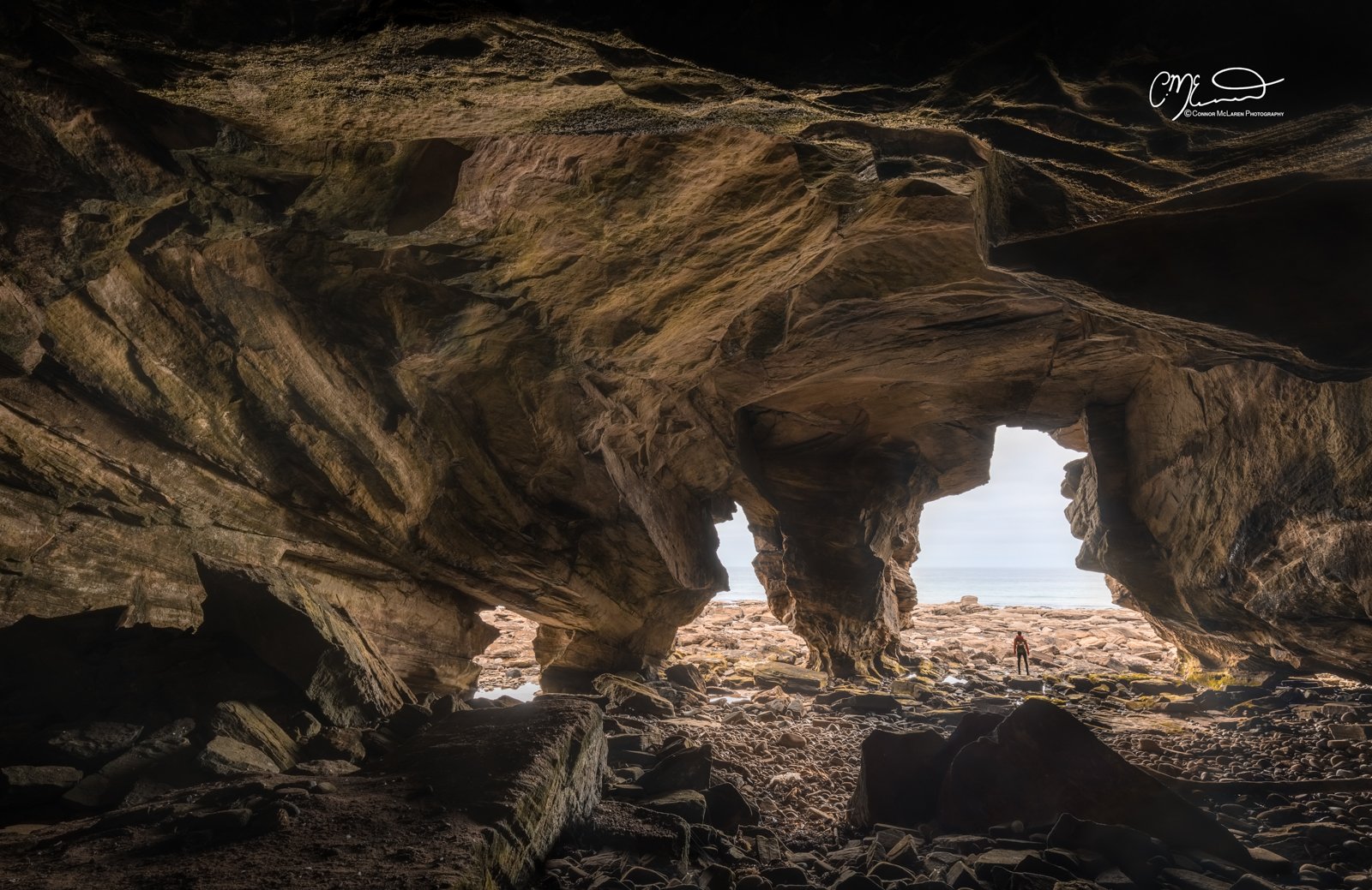Covesea Caves, North East Scotland. View from inside a rocky cave opening to the beach with a person standing on the rocks near the water, behind the cave entrance.