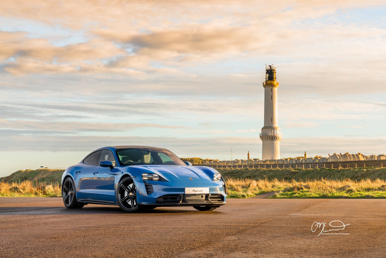 A blue Porsche Taycan parked on a sandy ground with a lighthouse and grassy landscape in the background during sunset.