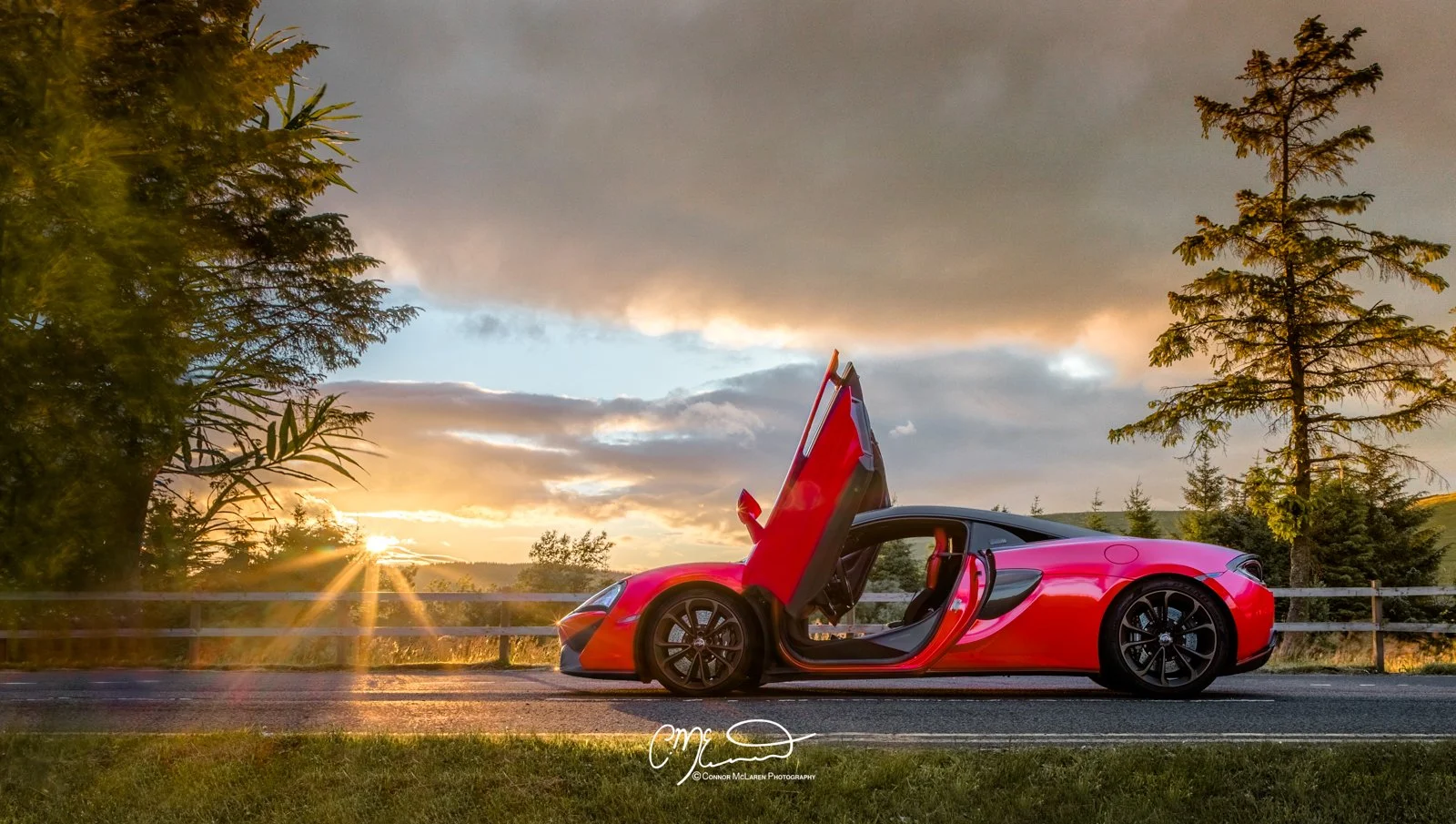 Red McLaren 540C sports car with gullwing door open parked on road at sunset, trees in the background.
