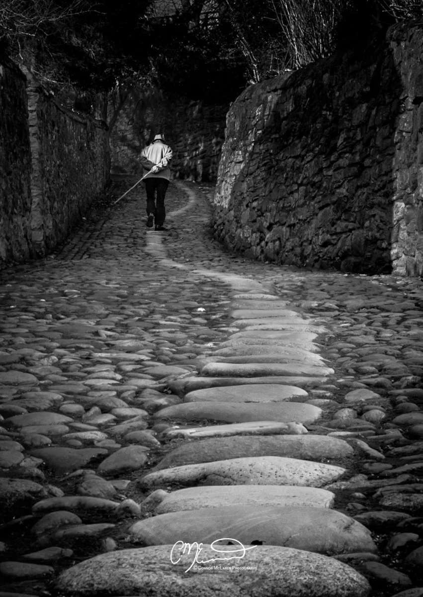 Scotland, old man with cane. A person walking down a cobblestone path between stone walls, holding a walking stick, in black and white.
