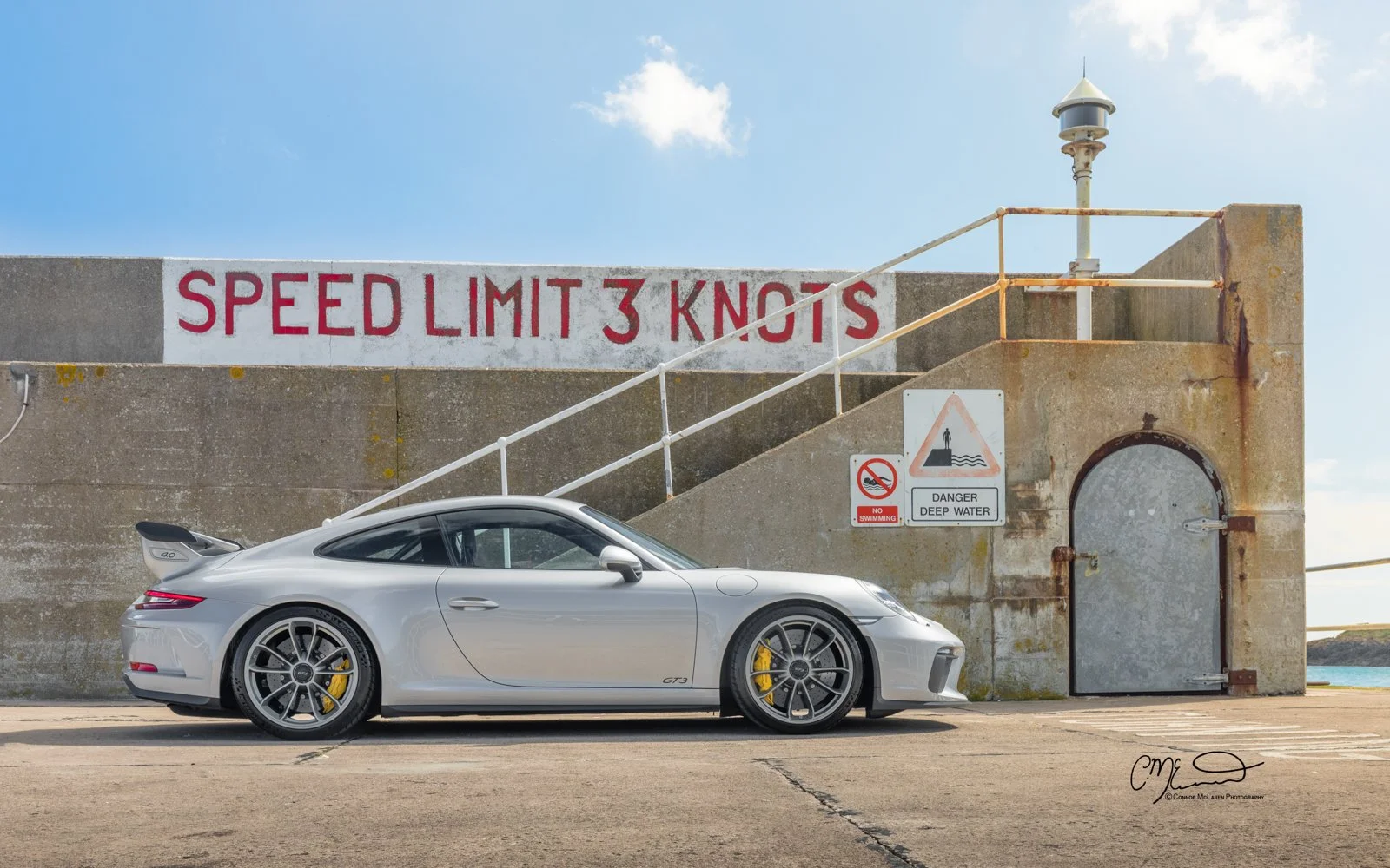 A silver Porsche GT3 sports car parked on a concrete surface near a weathered concrete structure with a sign that says 'Speed Limit 3 Knots' and another sign warning about deep water. The background shows a blue sky with some white clouds.