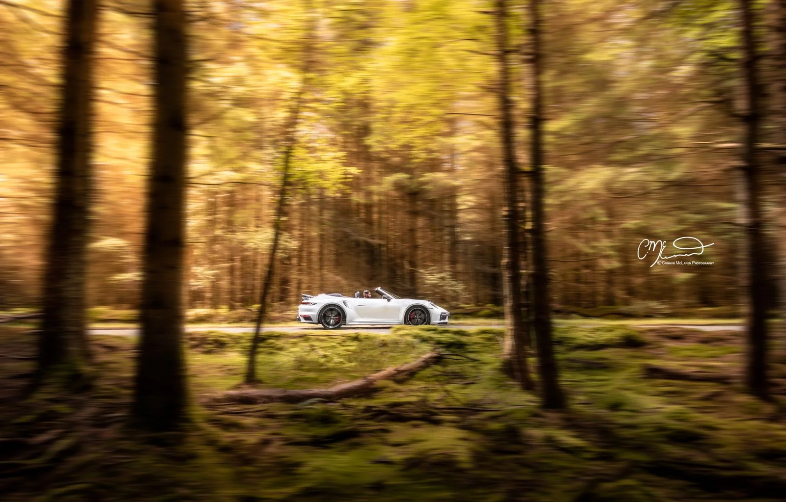 A white Porsche 911 convertible sports car driving on a road through a forest with autumn-colored trees.