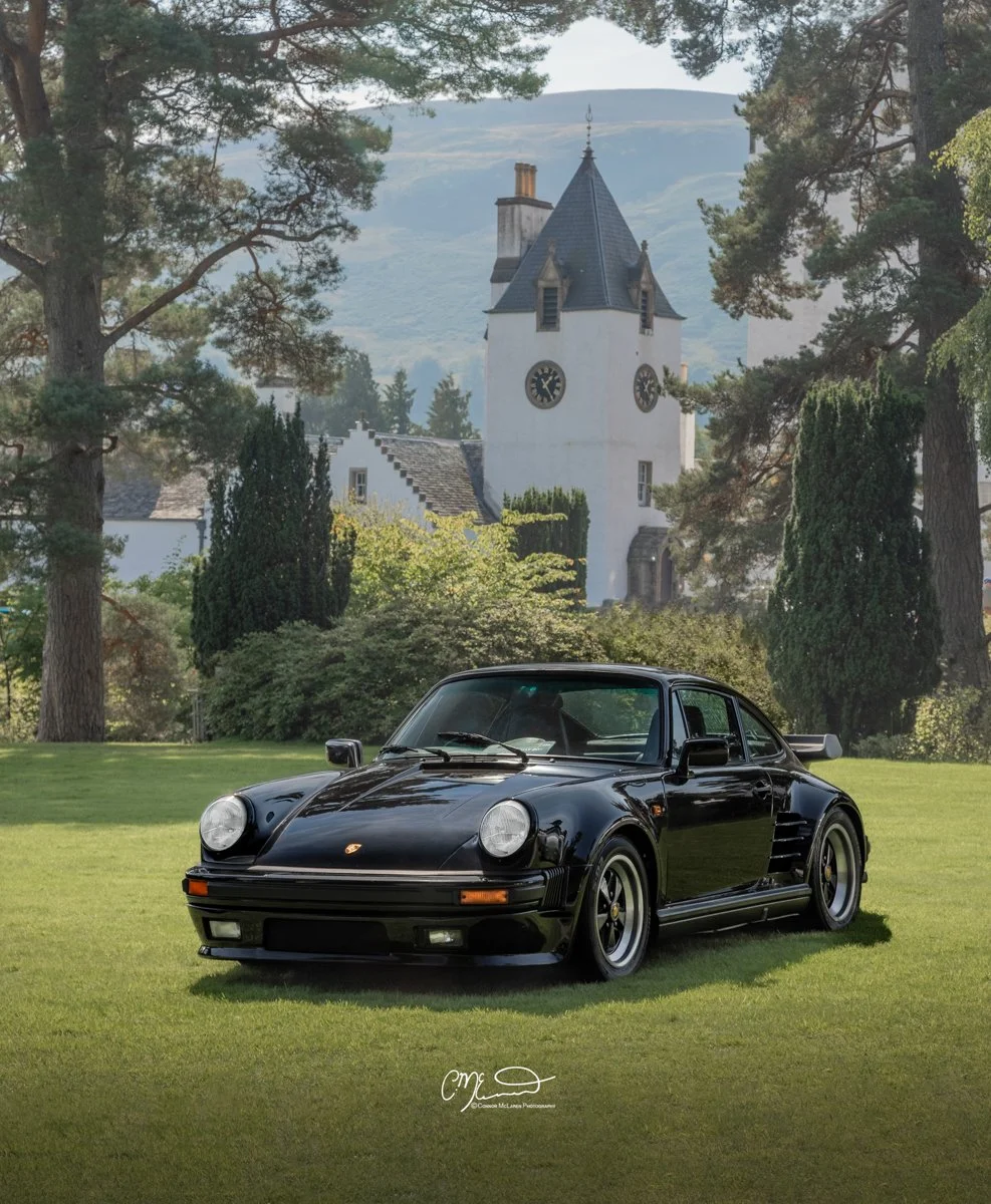 Porsche 911 930 Turbo. A black Porsche 911 sports car parked on a well-kept lawn in front of a white church with a tall steeple and clocks, surrounded by trees, with a mountain in the background.