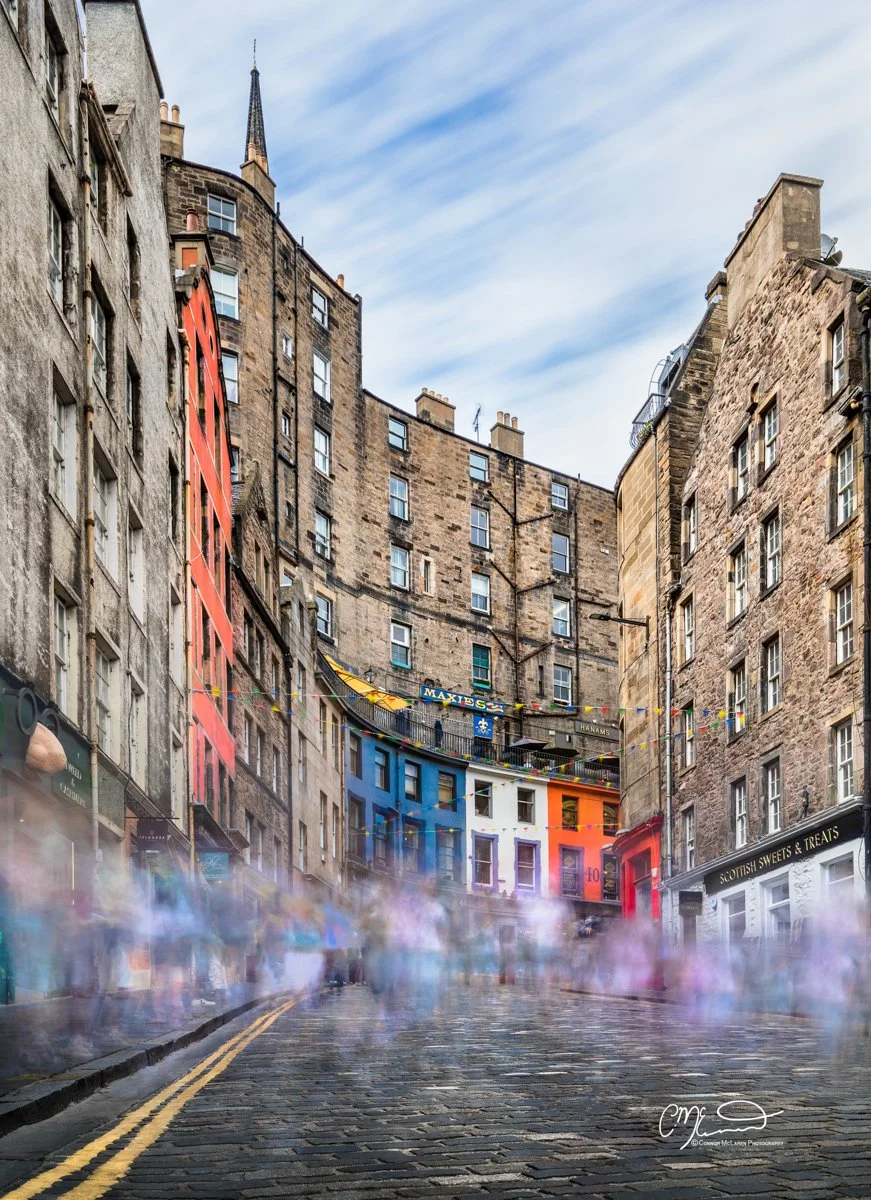 A cobblestone street in a historic city neighborhood with tall, old buildings on both sides. There are colorful decorations and banners hanging across the street, with a blue, orange, and white building at the end. The street appears misty with motion-blurred figures, indicating a lively atmosphere.