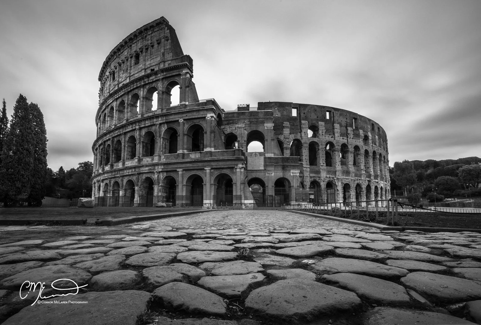 Italy, Rome, Colosseum. Black and white photo of the Roman Colosseum, showing its arches and partial damaged structure, with a cobblestone path in the foreground and cloudy sky overhead.