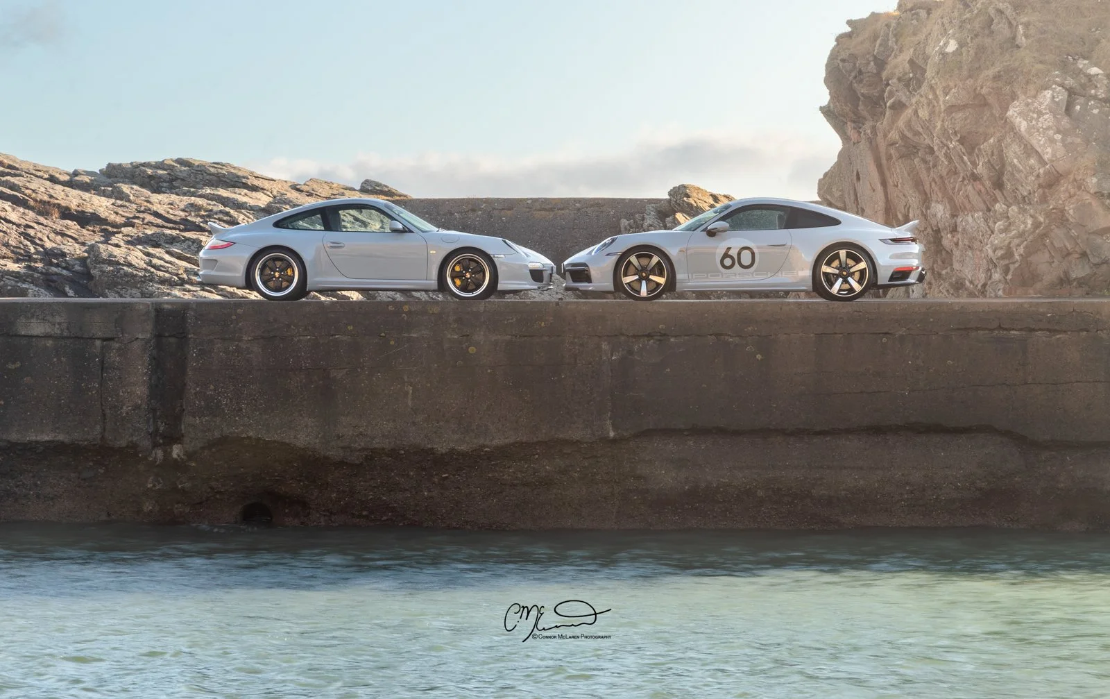 Porsche 911 Sport Classic. Two white Porsche sports cars parked on a concrete pier by the water, with rocks and cliffs in the background under a partly cloudy sky.