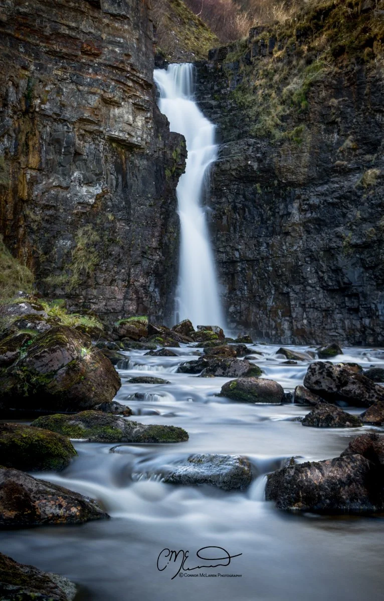 Scotland, Isle of Skye. Tall waterfall cascading down dark rocky cliffs into a river with moss-covered rocks in the foreground.
