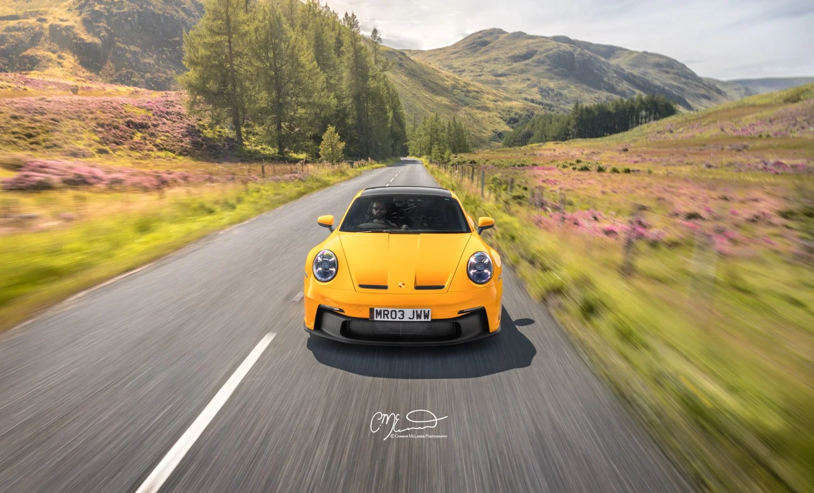 Mr JWW Porsche 911 GT3 PTS Speed yellow. A yellow sports car driving down a rural road through a landscape with green hills and pink flowers, with a mountain in the background.