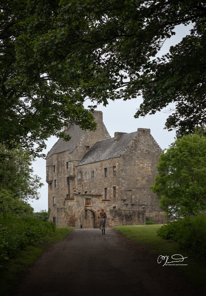 Scottish Lowlands, Scotland, midhope castle, lalybroch Outlander filming location. A woman standing in front of a historic stone castle surrounded by green trees, with a dirt path leading to the castle entrance.