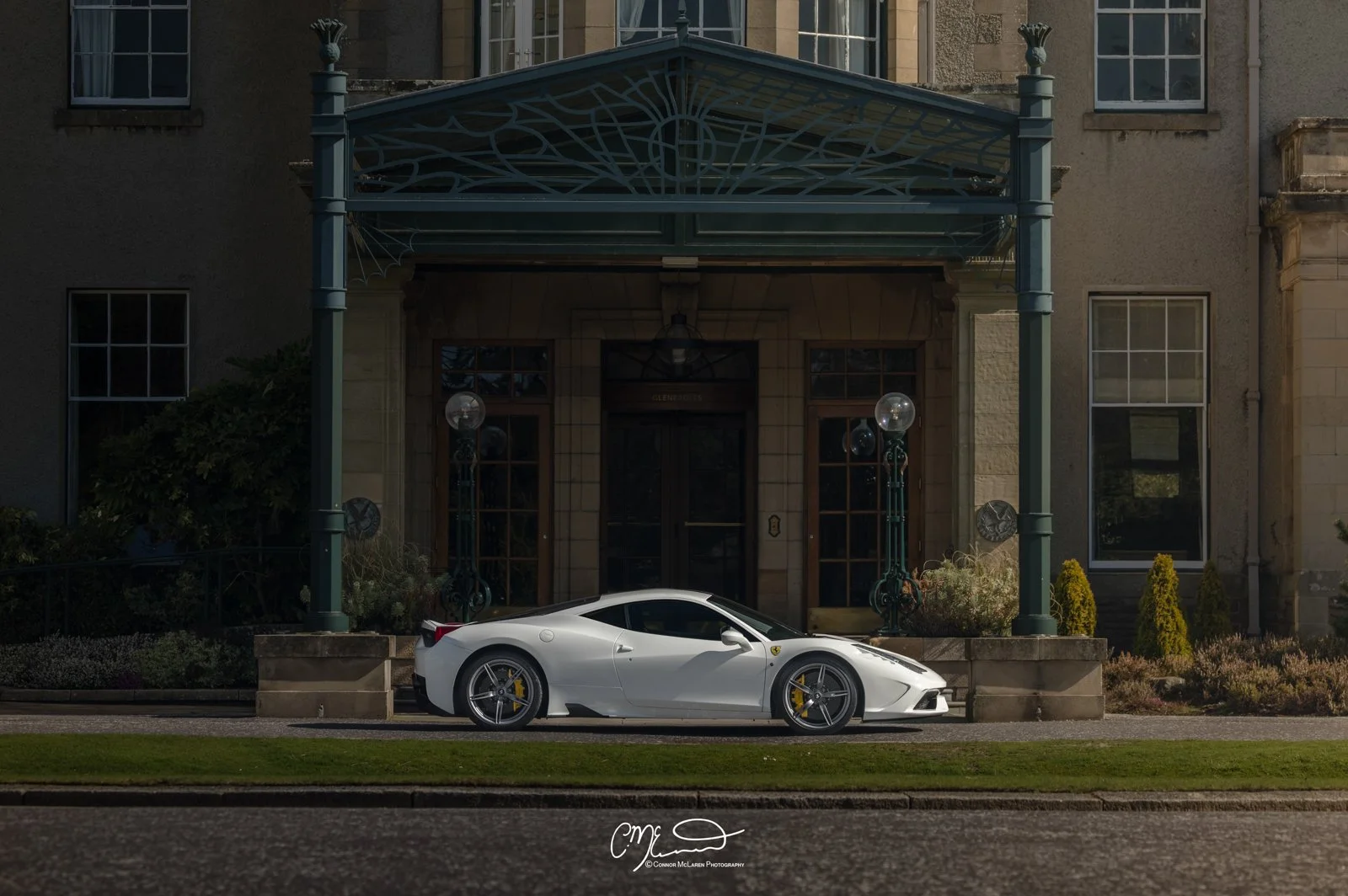 Ferrari 458 Speciale in white at Gleneagles Hotel. White sports car parked in front of a historic building with brick walls, large windows, and a decorative metal awning, during daylight.