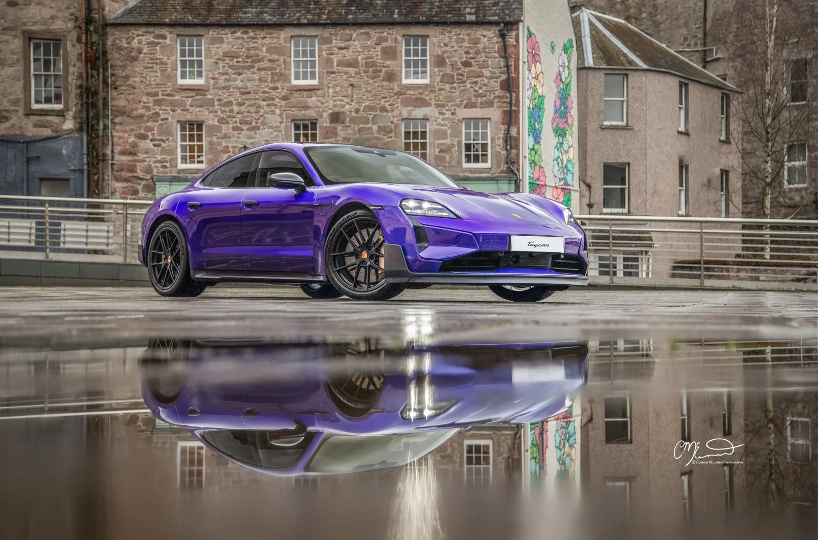 A purple Porsche Taycan Turbo GT parked on a rooftop with its reflection visible on a wet surface, urban buildings and a mural with colorful flowers in the background.