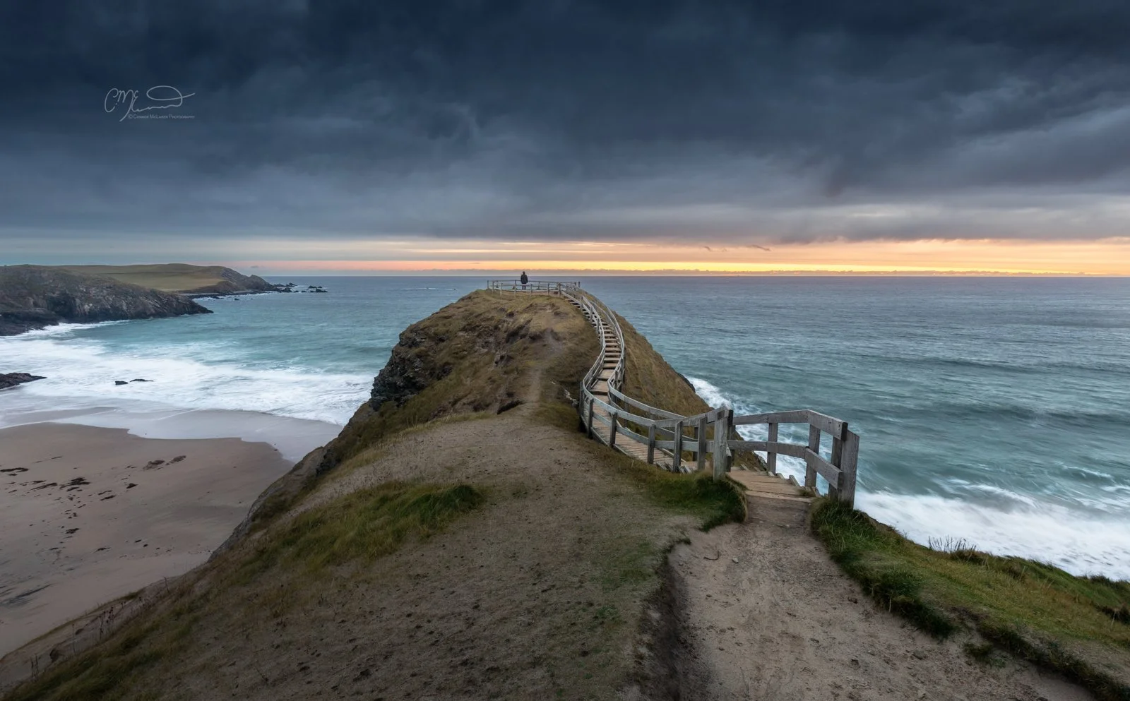 Scotland, NC500 Durness. A narrow grassy trail on a coastal headland with a wooden railing running along the edge, leading toward a viewpoint at the tip of the headland overlooking the ocean during sunset, with dark clouds overhead.