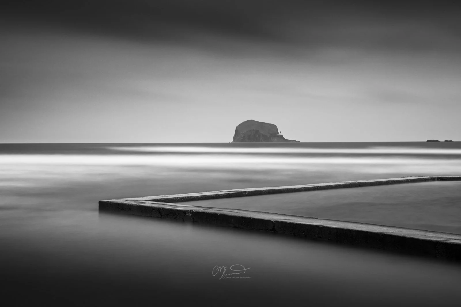 Scotland, Bass rock, North Berwick. Black and white long exposure photograph of a seascape with a large rock formation in the distance, smooth water, and a concrete edge in the foreground.