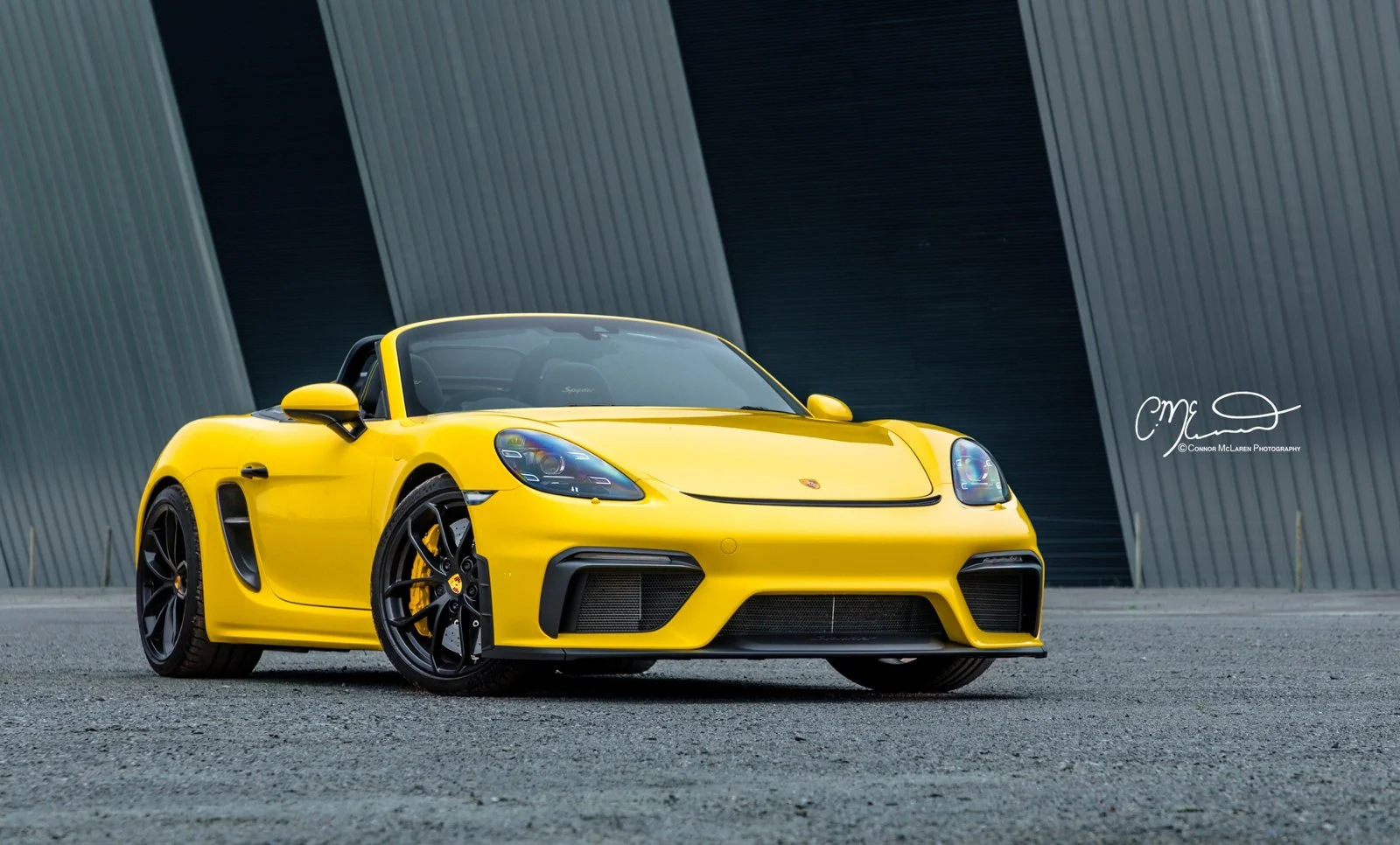 Yellow Porsche Spyder sports car parked on asphalt in front of modern metallic building.