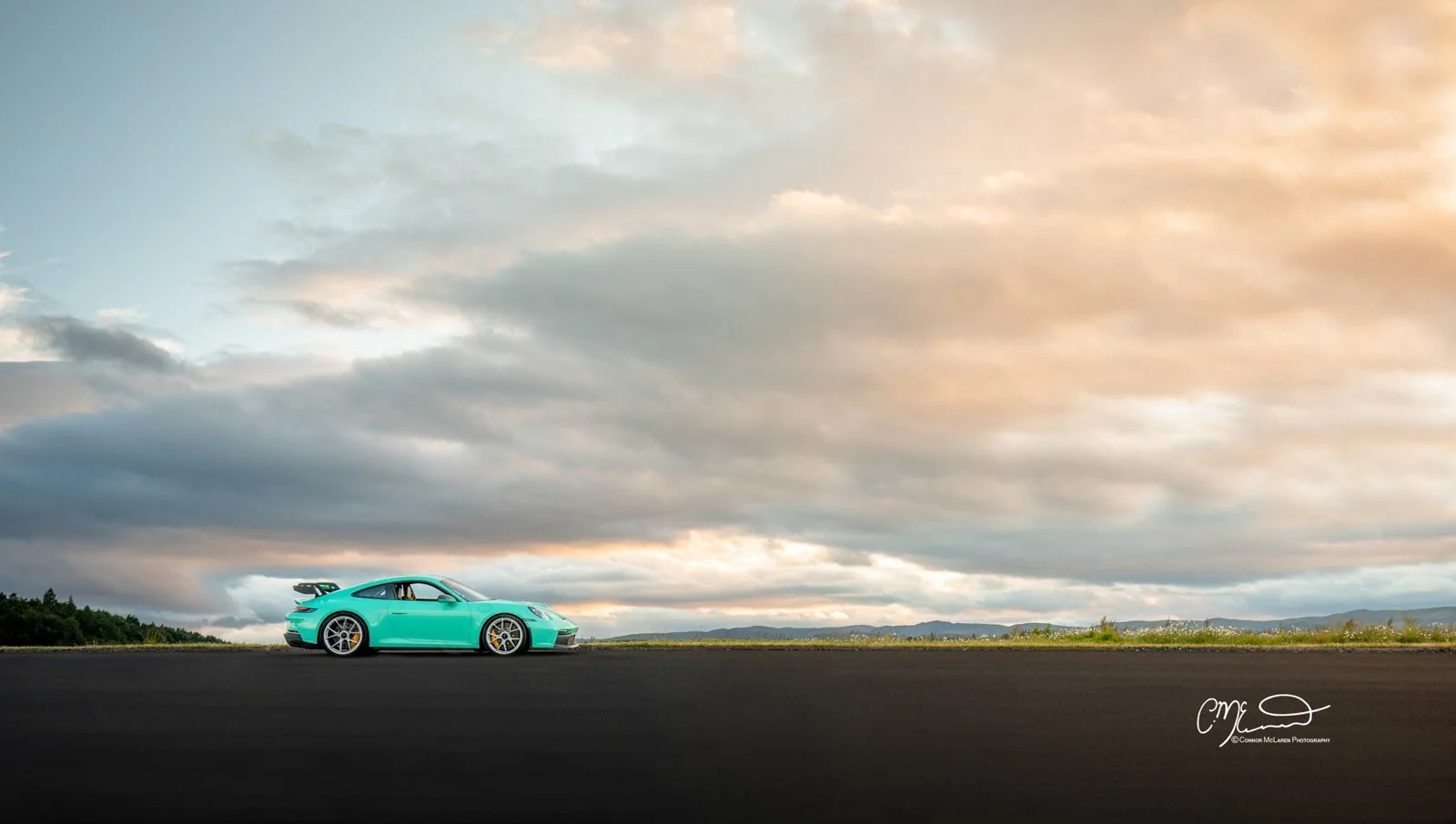 Mint Porsche 911 GT3 992. A turquoise sports car with a large rear spoiler parked on a paved road under a cloudy sky at sunset or sunrise.