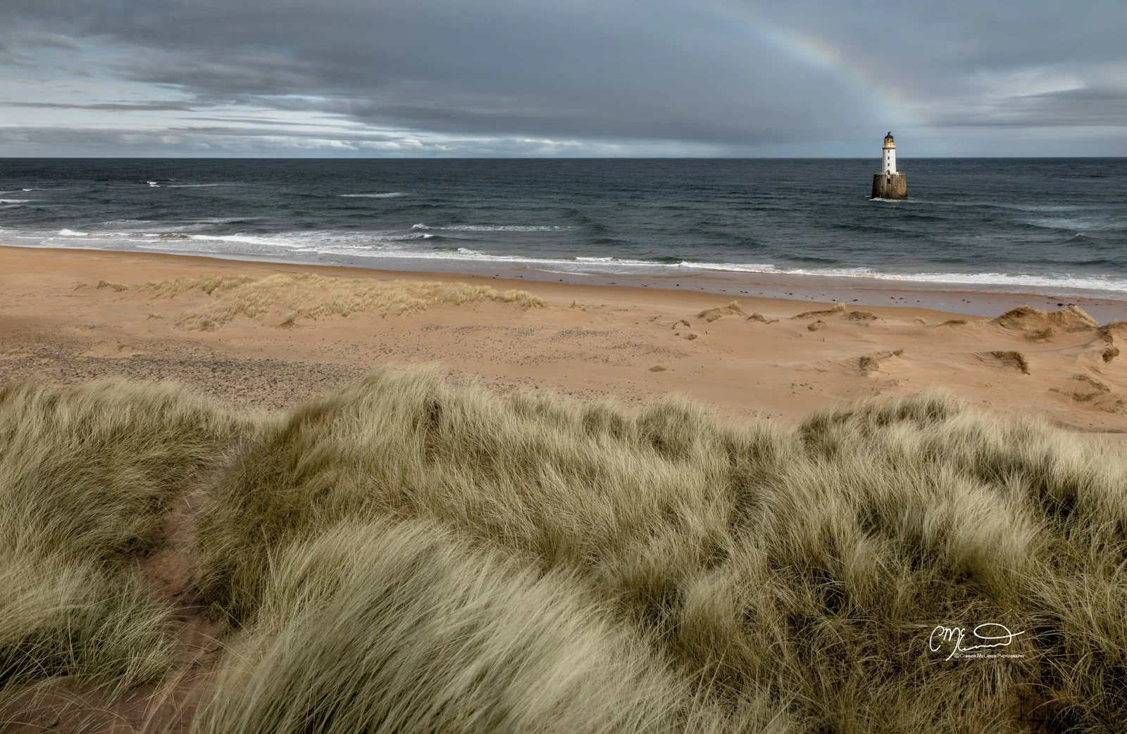 rattray head lighthouse, rainbow, North East Scotland. Sand dunes with grass near a beach, with the ocean and a lighthouse on a rock in the distance, and a rainbow in the cloudy sky.