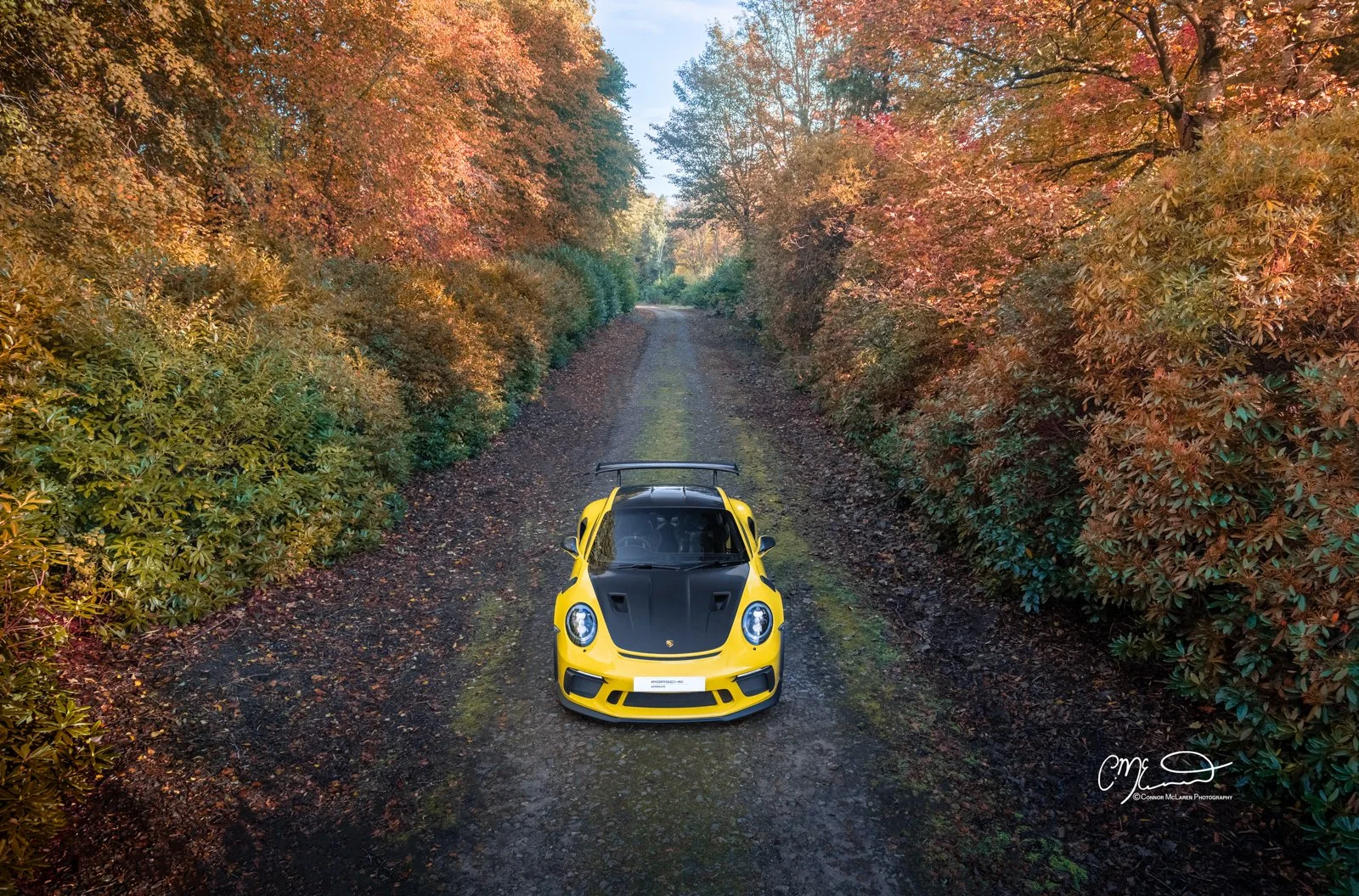 A yellow Porsche 911 GT3 RS Weissach sports car with black accents parked on a dirt path surrounded by colorful autumn trees.