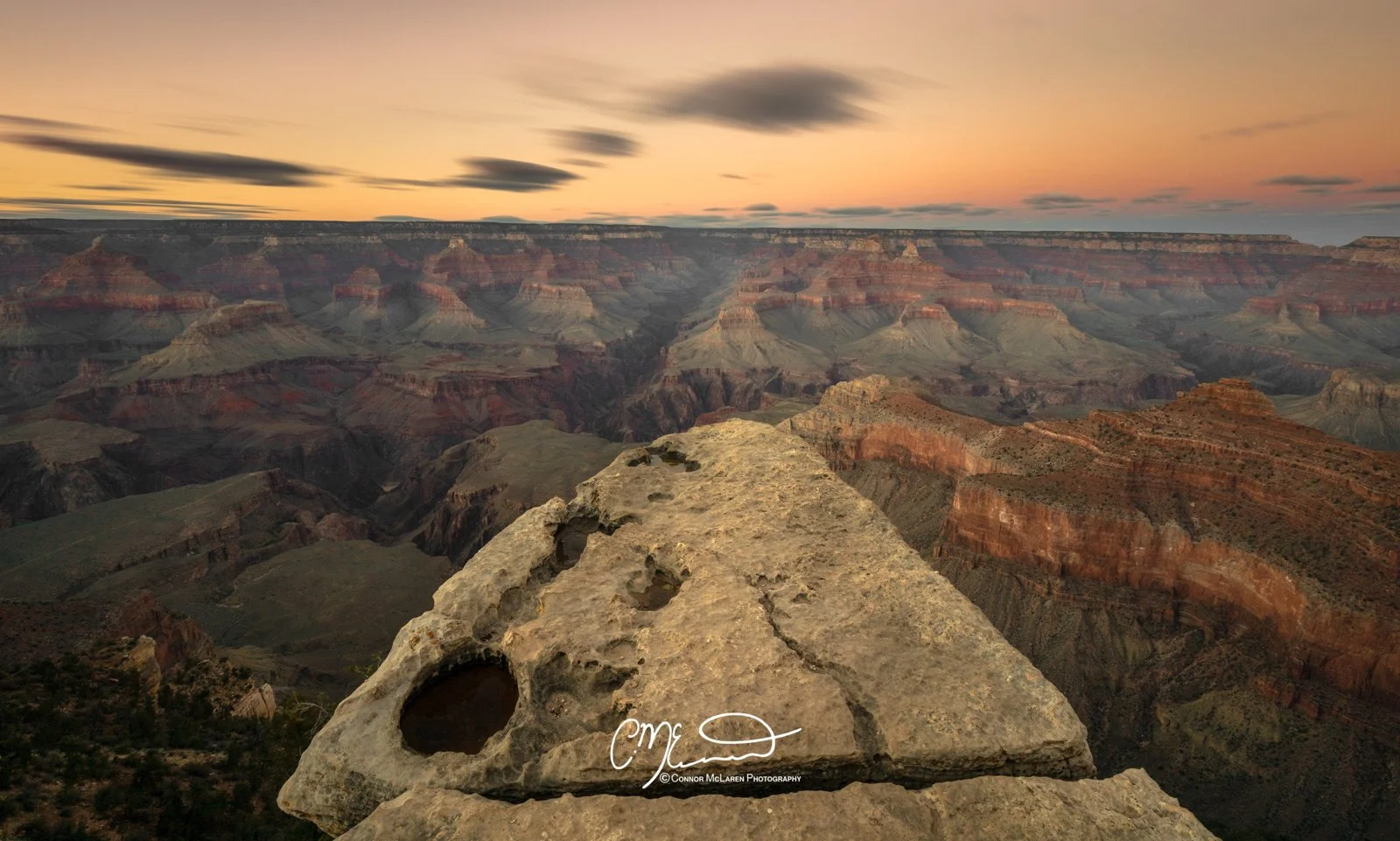 Bucket list locations, USA Grand Canyon. View of the Grand Canyon at sunset with layered rock formations and a cloudy sky, seen from a stone ledge.