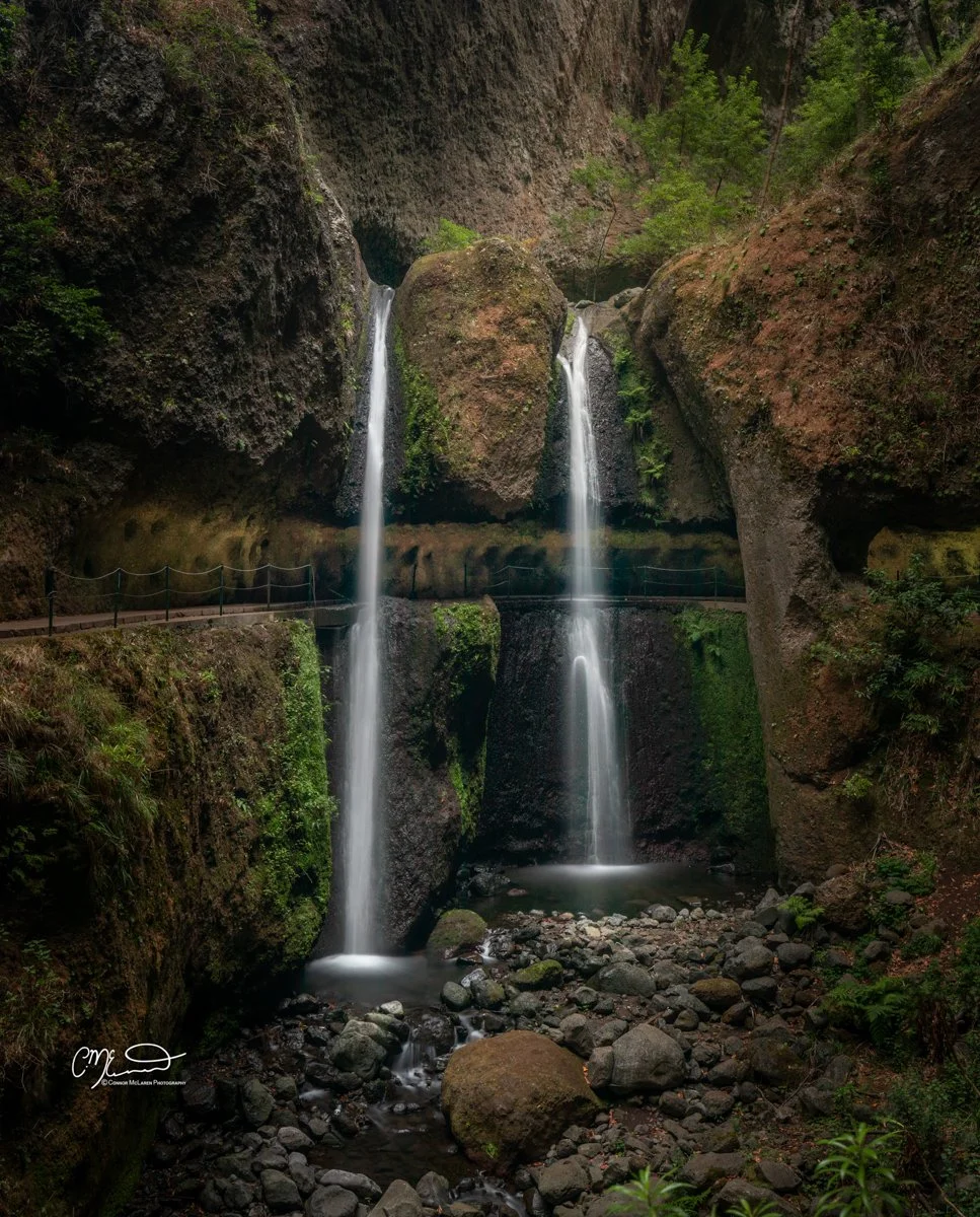 Levada nova, Madeira, Portugal. A double waterfall flowing down large rocks in a lush green canyon with a walkway and railing along the side.