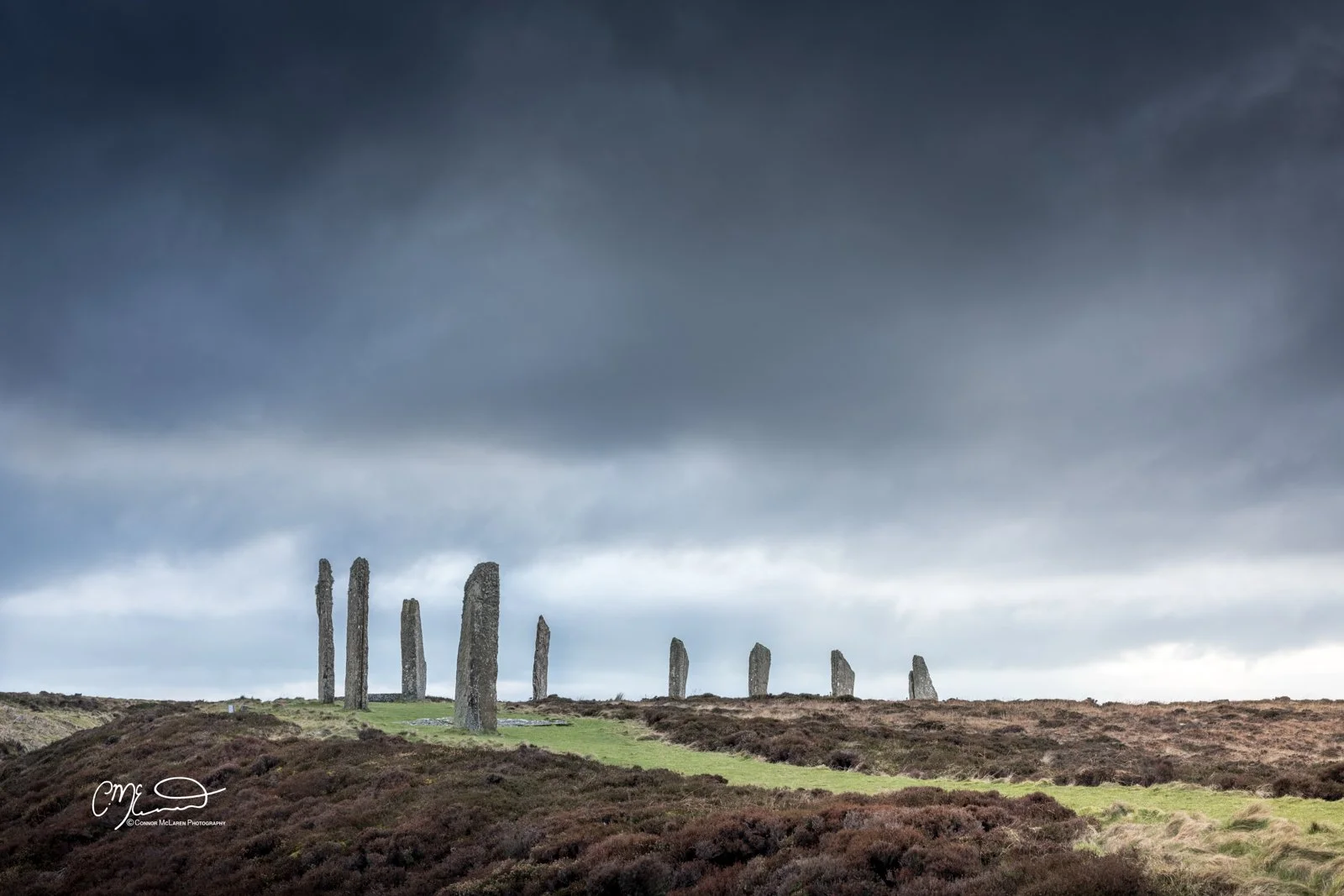Ring of Brodgar, Orkney, Scotland. Ancient stone circle on a hill under a dark, cloudy sky with patches of green grass and brown shrubbery in the foreground.