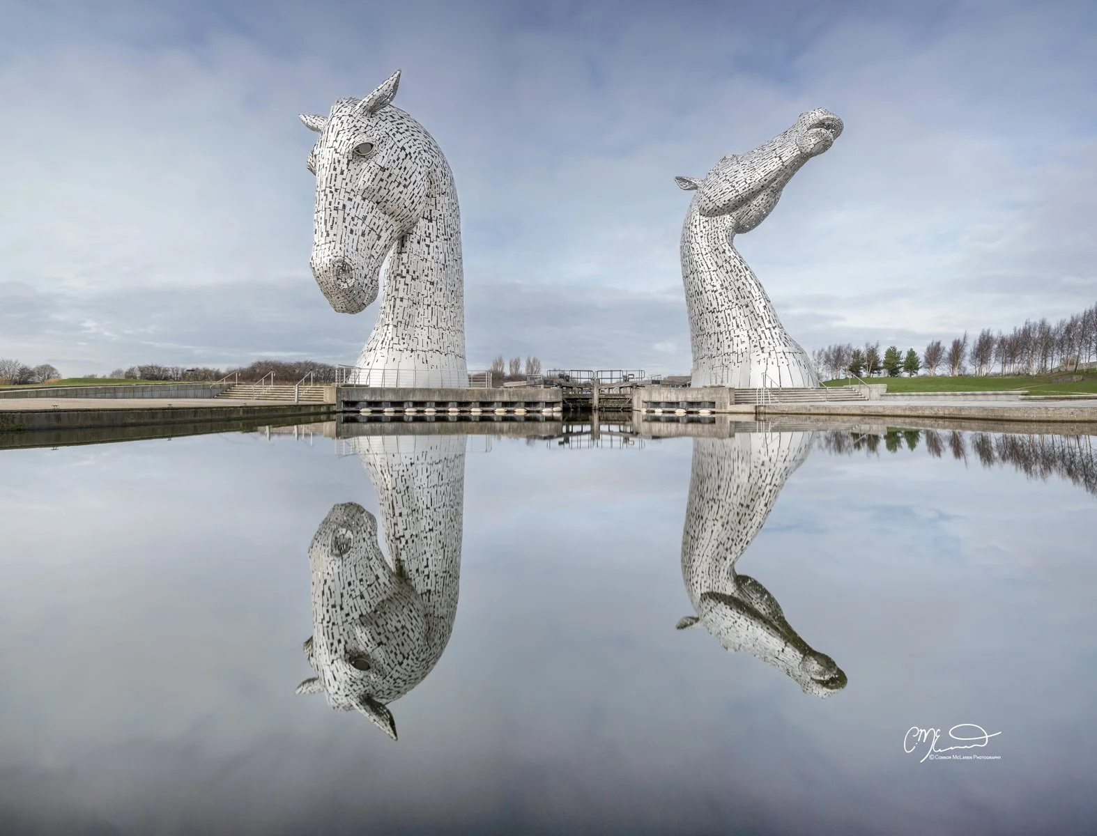 Scottish Lowlands, Scotland, Kelpies. Two large metallic horse sculptures reflecting in the water at the Kelpies in Scotland, with a cloudy sky above.