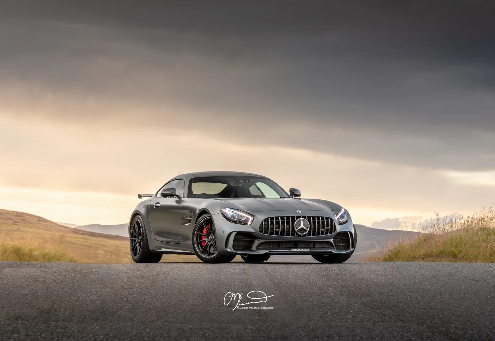 Gray Mercedes-Benz AMG GT sports car parked on an empty road with a cloudy sky and hills in the background.