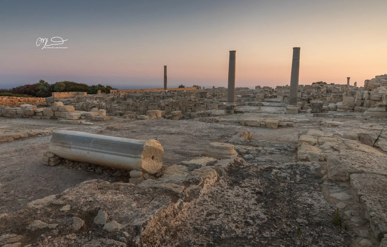 kourion, Cyprus. Ancient archaeological site with scattered stone ruins and columns under a pastel sunset sky.